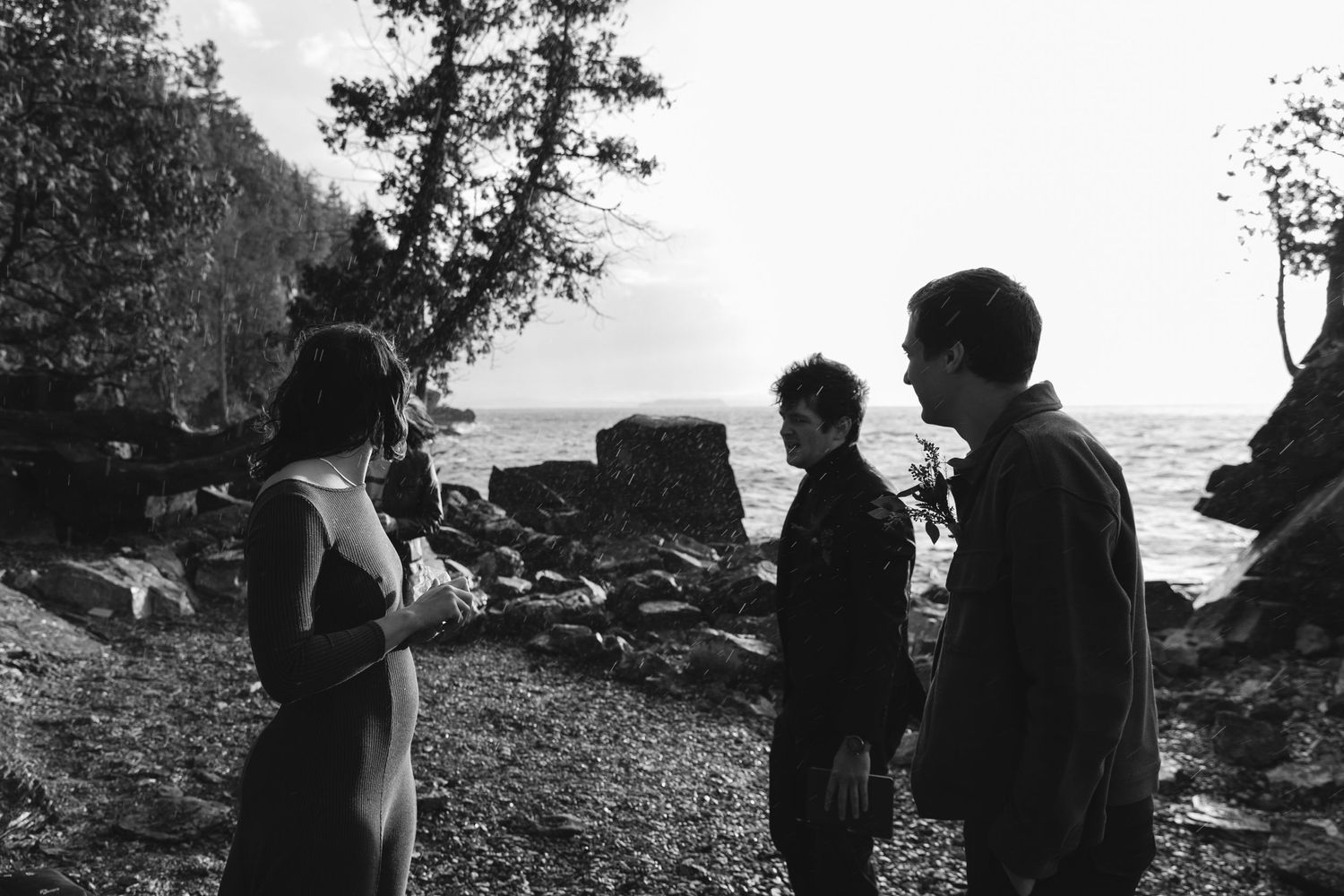 Three people gather on a rocky beach at sunset surrounded by natural coastal scenery.