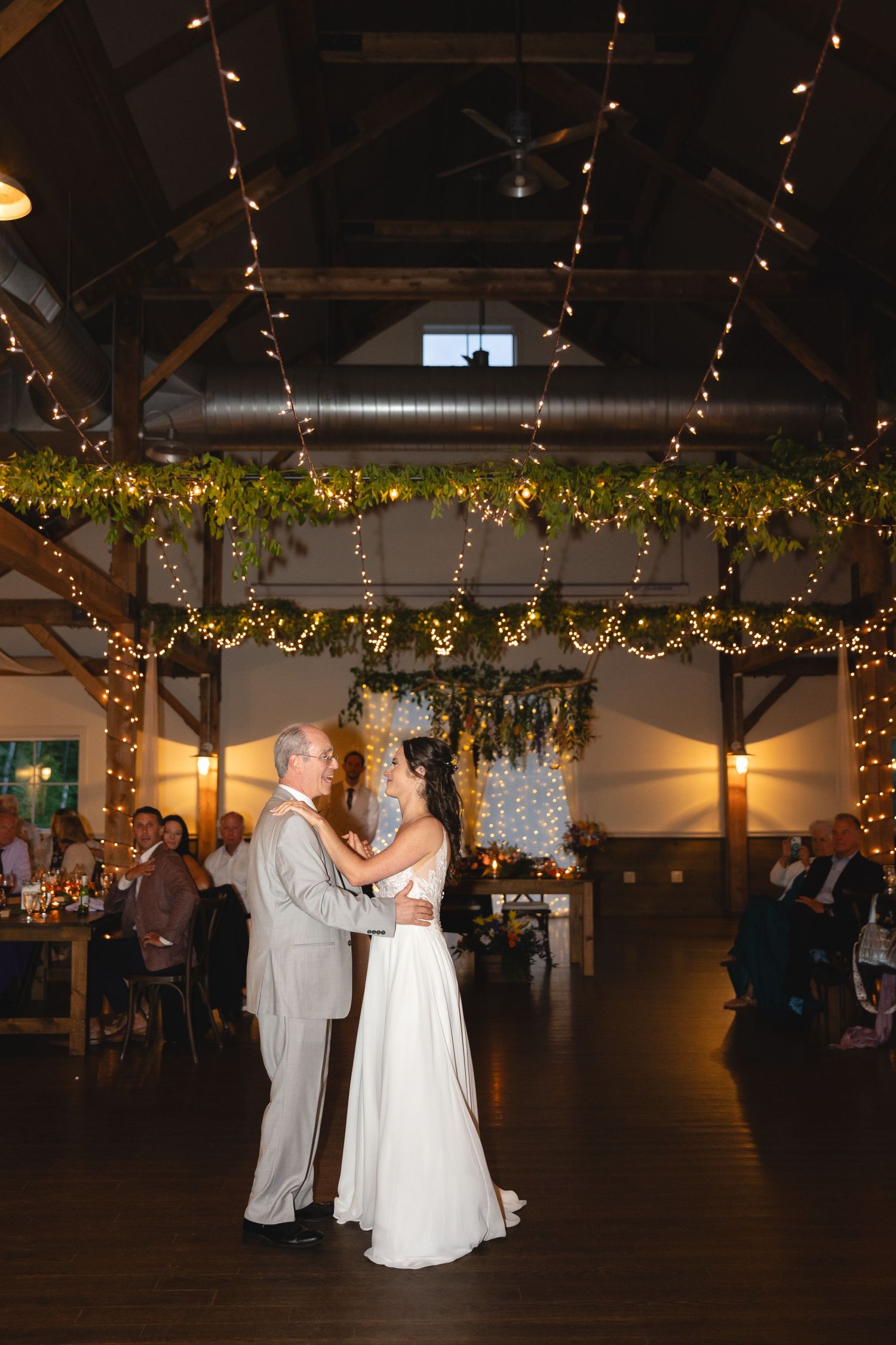 A couple dances beneath twinkling lights and greenery in a rustic reception venue.
