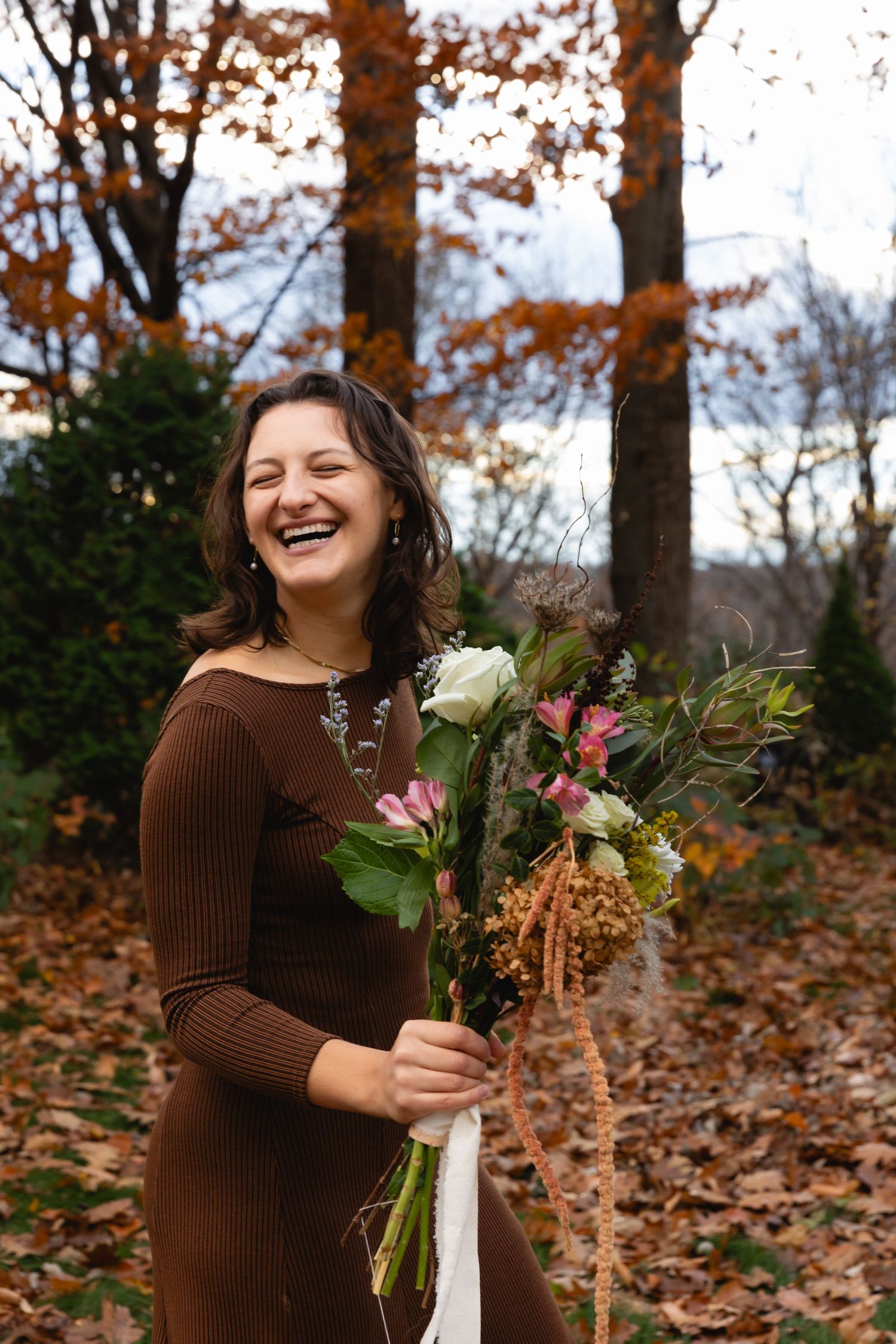 Someone in a brown sweater holds a bouquet of mixed flowers while standing in an autumn forest with fallen leaves.