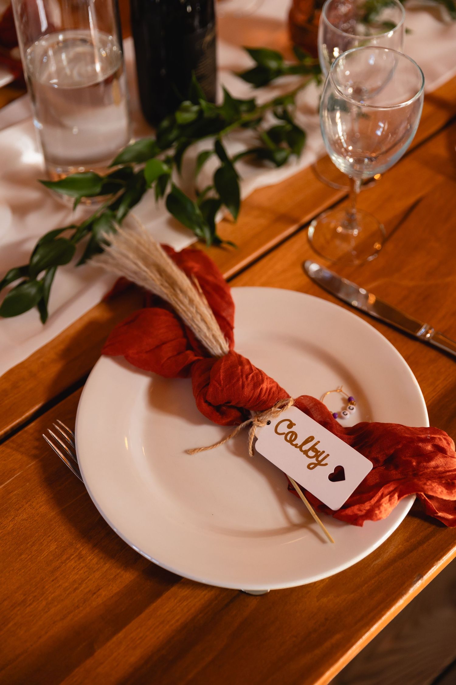 Elegant table setting with a red ribbon place card on a white plate with wine glasses and greenery on wooden table.