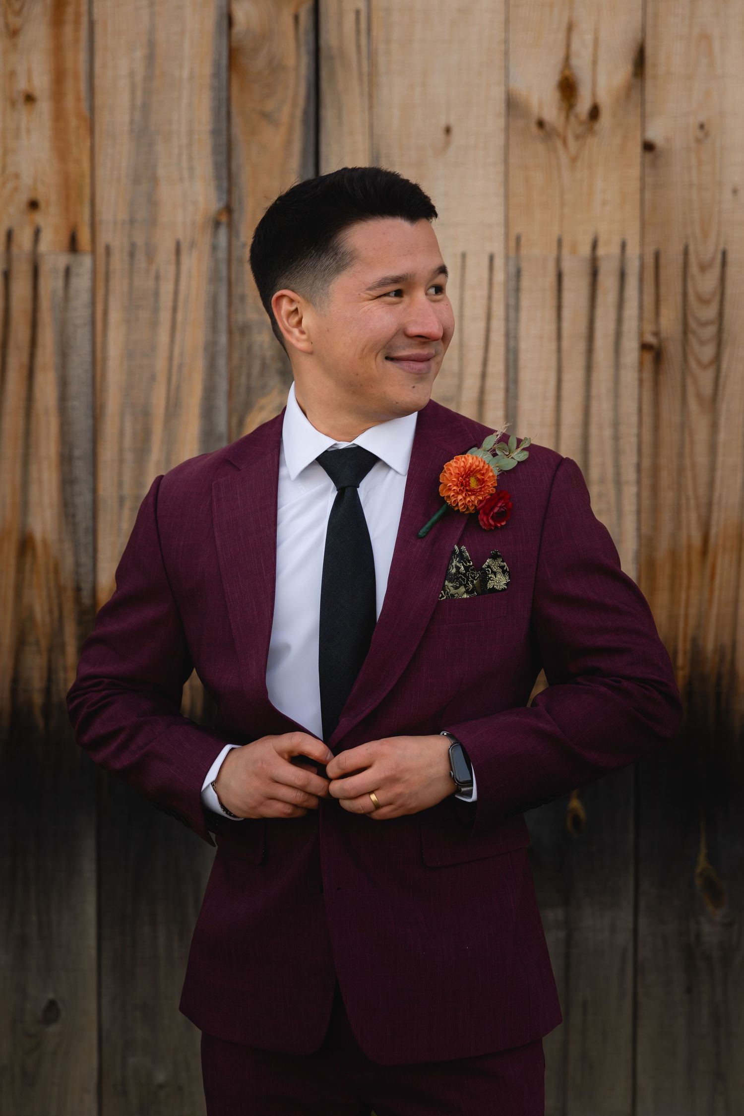 A groom in a burgundy suit and black tie smiles while adjusting his jacket in front of a wooden fence.