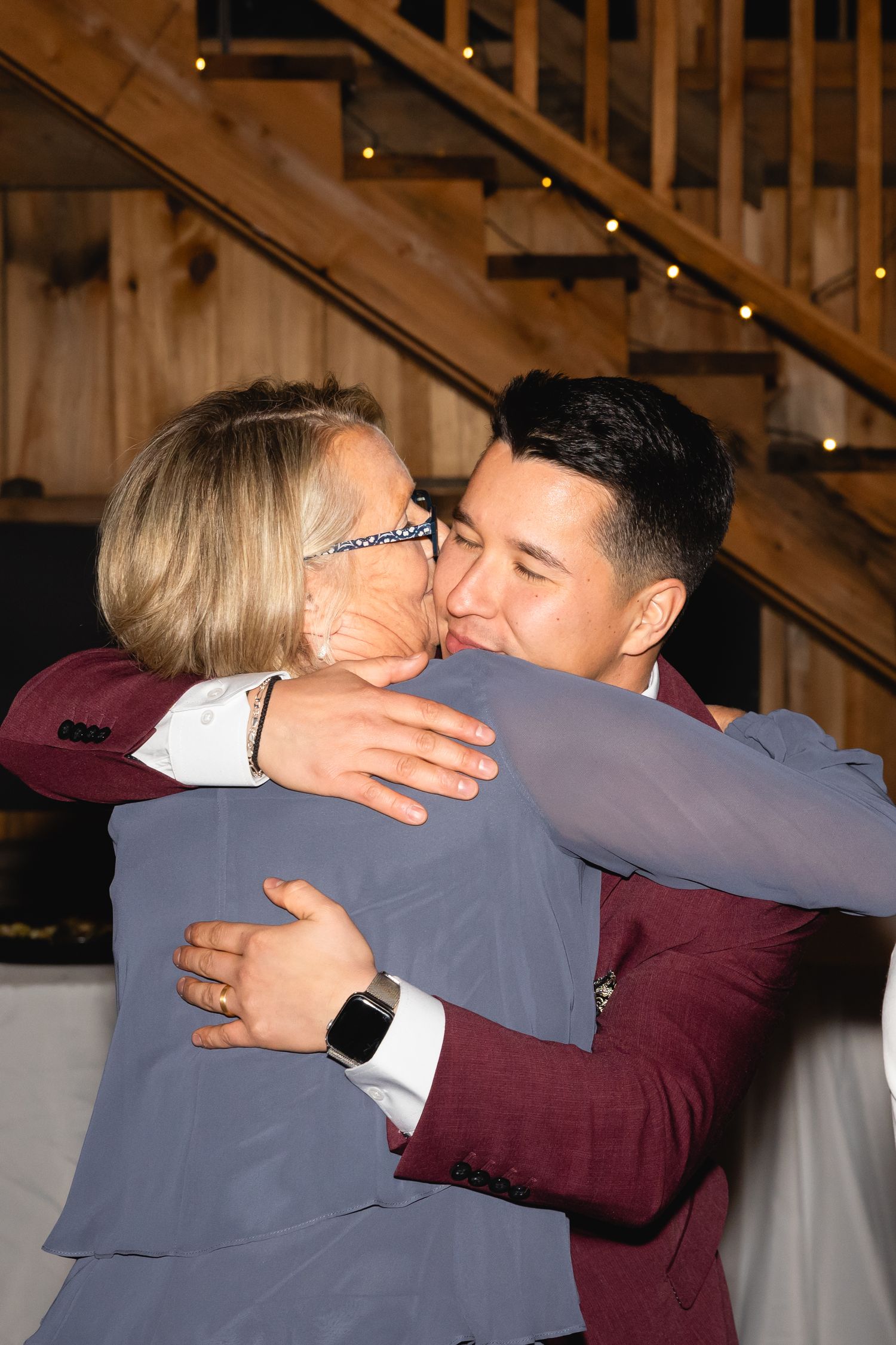 Two people sharing a warm embrace at what appears to be an indoor event venue with wooden stairs in the background.