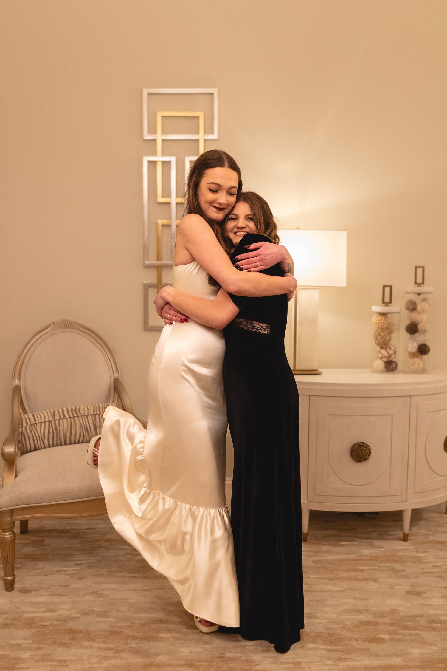 Two figures embrace in a warm hug, one wearing an elegant white gown with a ruffle hem and one in a long black dress.