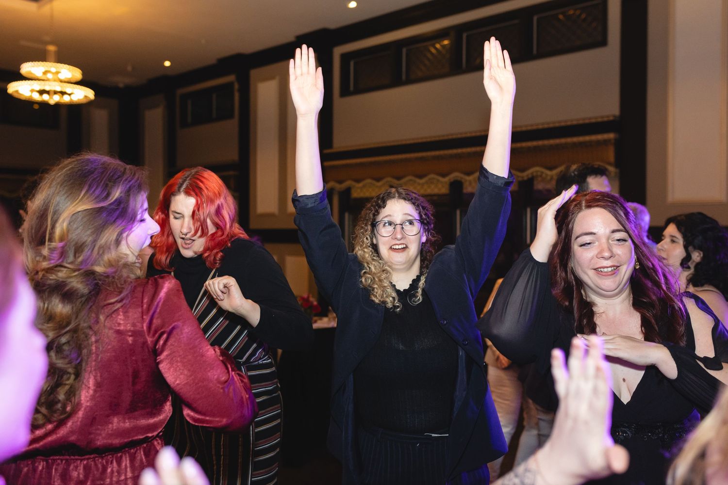 People dancing with raised arms at what appears to be a celebratory event in a dimly lit indoor venue with chandeliers.