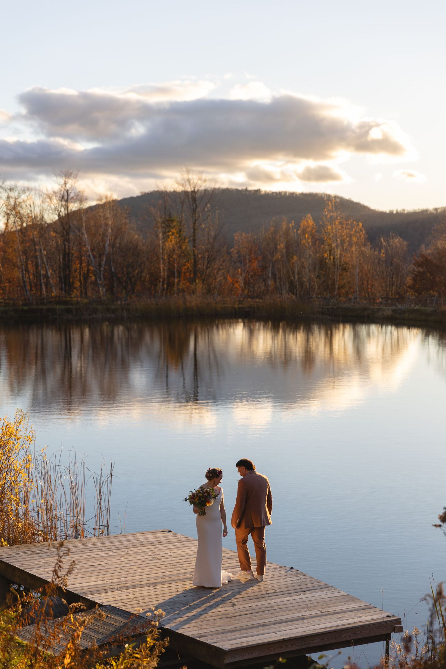 Couple stands on wooden dock overlooking serene lake at sunset with fall foliage.