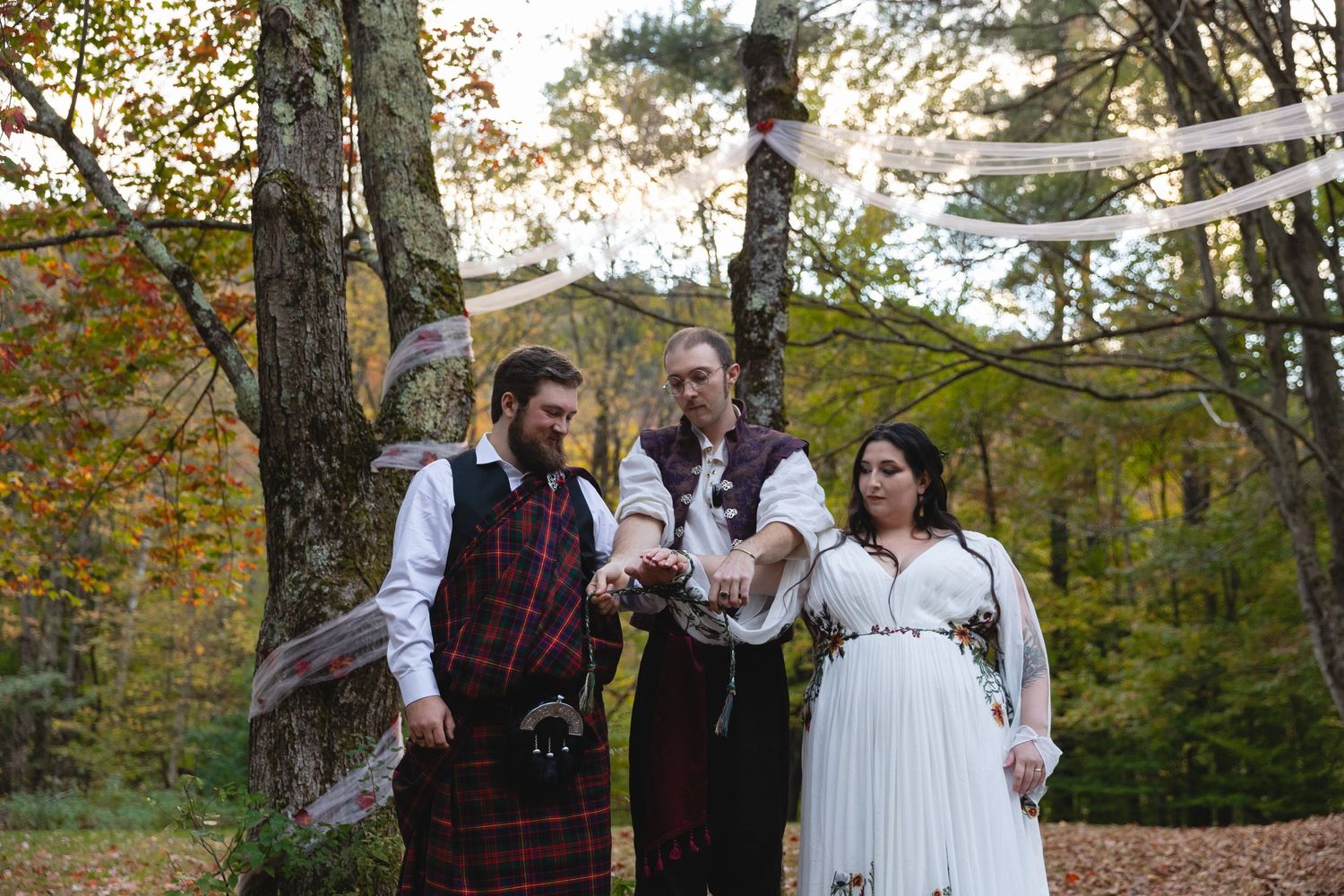 A wedding ceremony group stands under white draped fabric in an autumn forest setting.