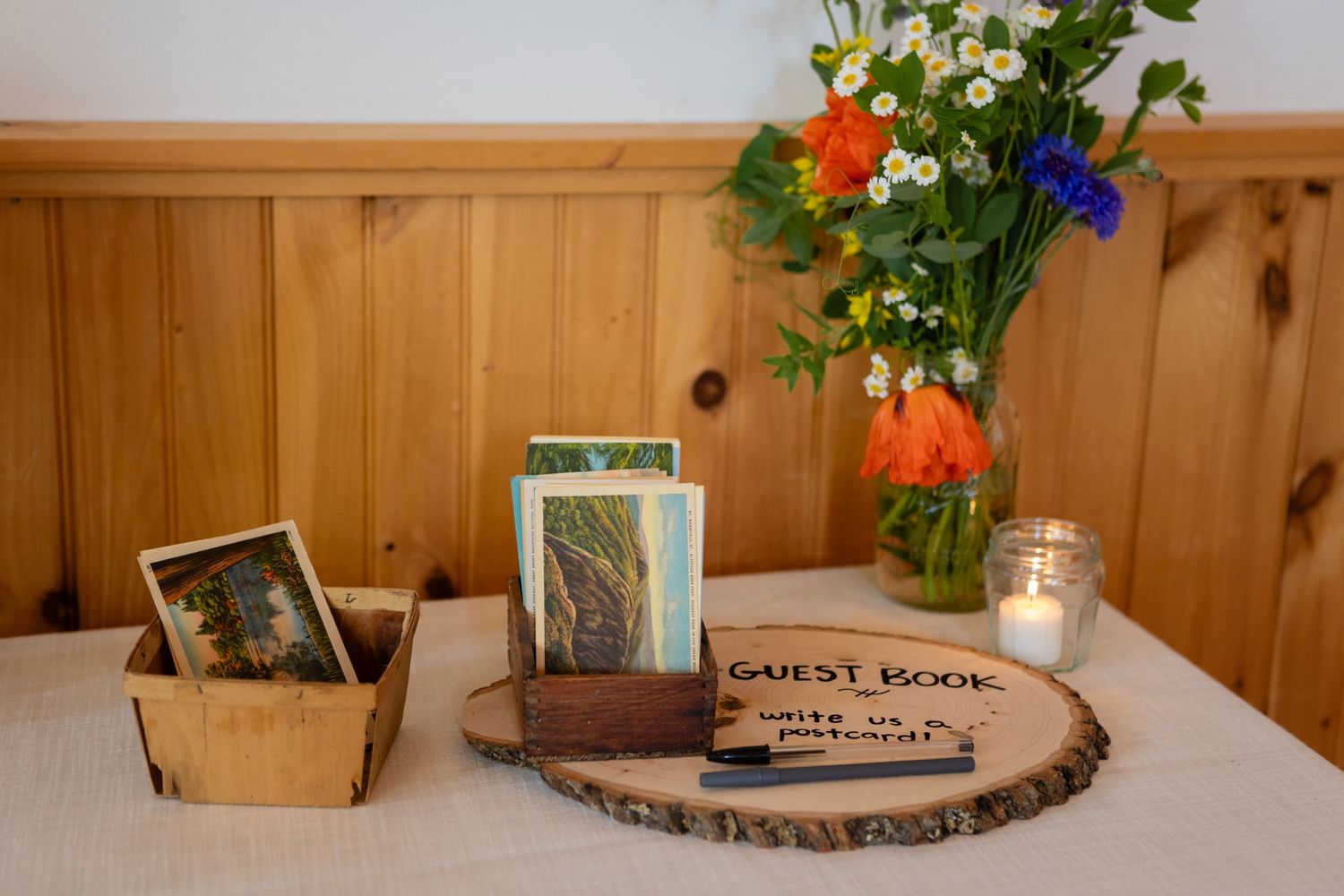 A rustic table display with a guest book, photos, and colorful wildflowers in a vase against wooden paneling.