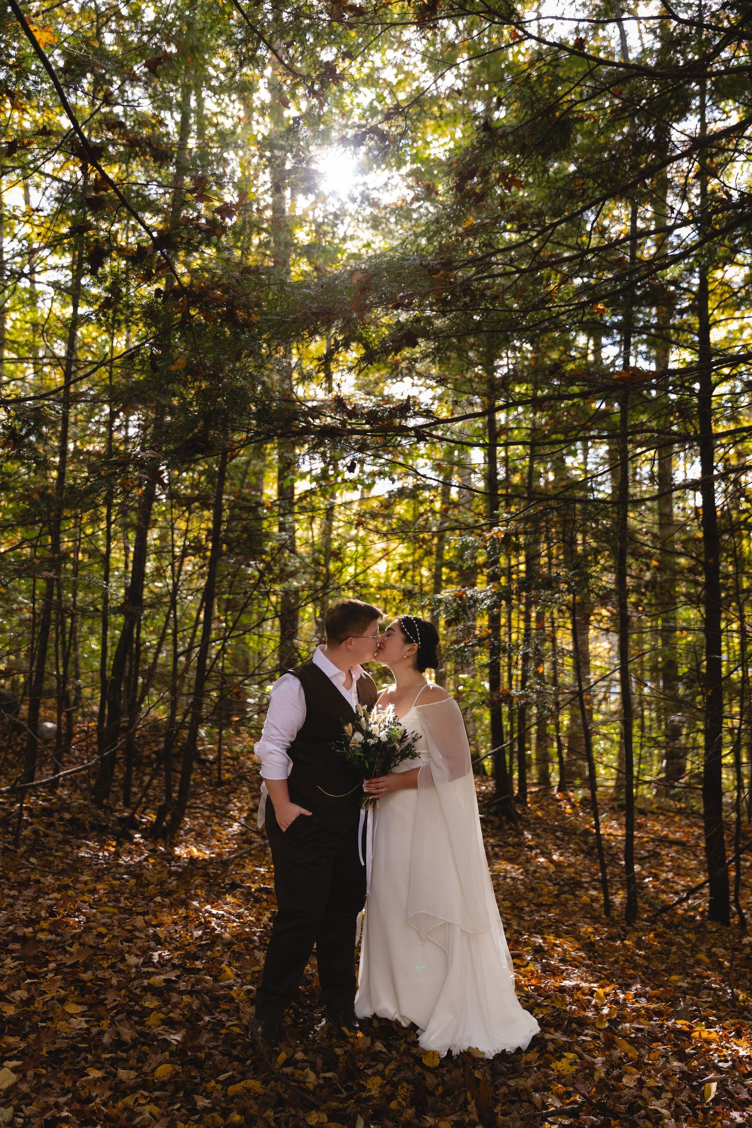 Newlyweds share an intimate moment in a sun-dappled forest with golden fall leaves.