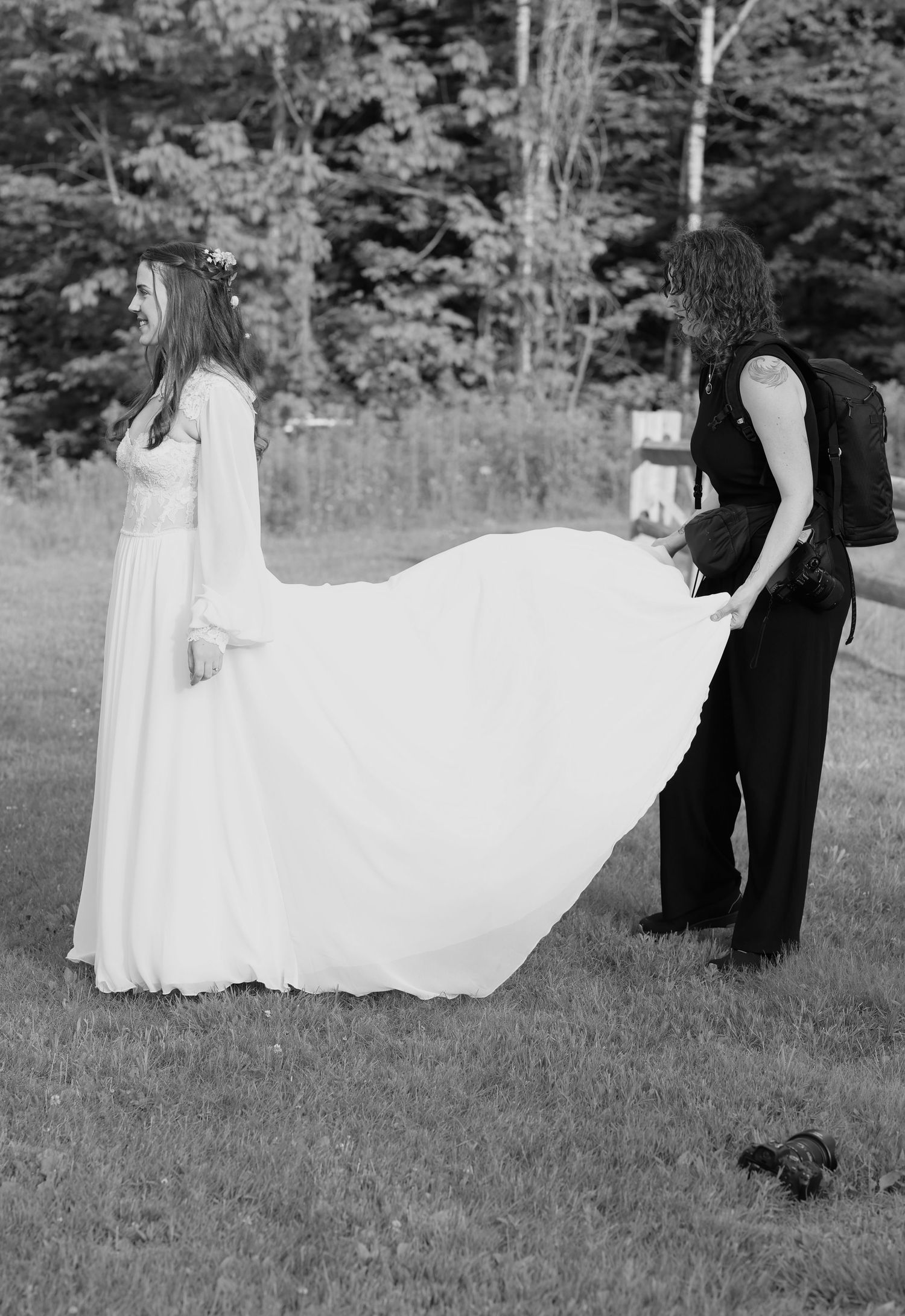 Black and white photo of wedding photographer fluffing bride's dress train outside.