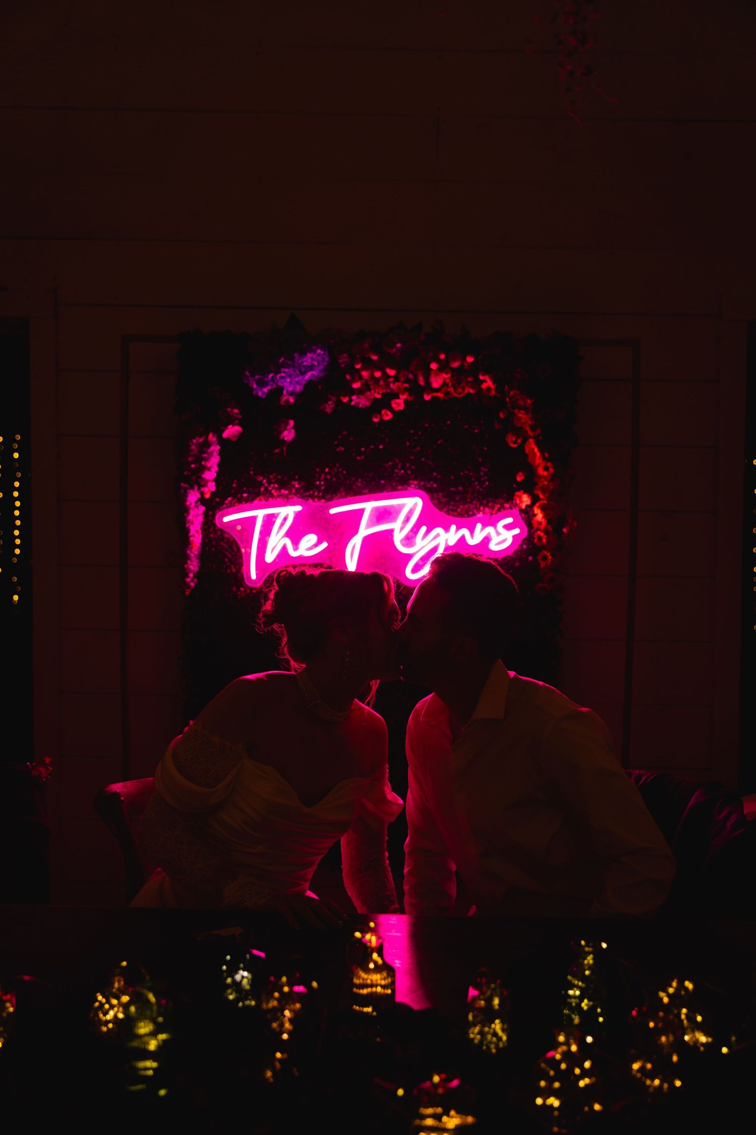 Couple sharing a kiss under a glowing pink neon sign reading The Flynns at their wedding reception.