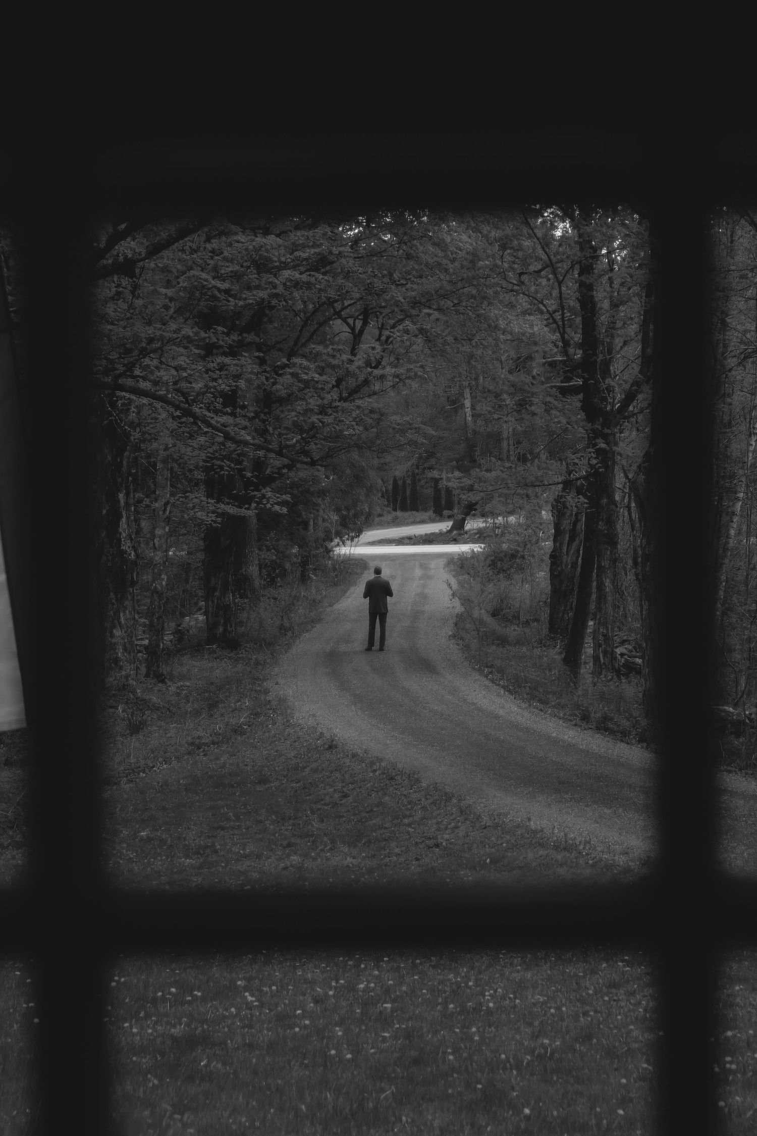 Black and white view of a lone figure standing on a winding forest road through a window.