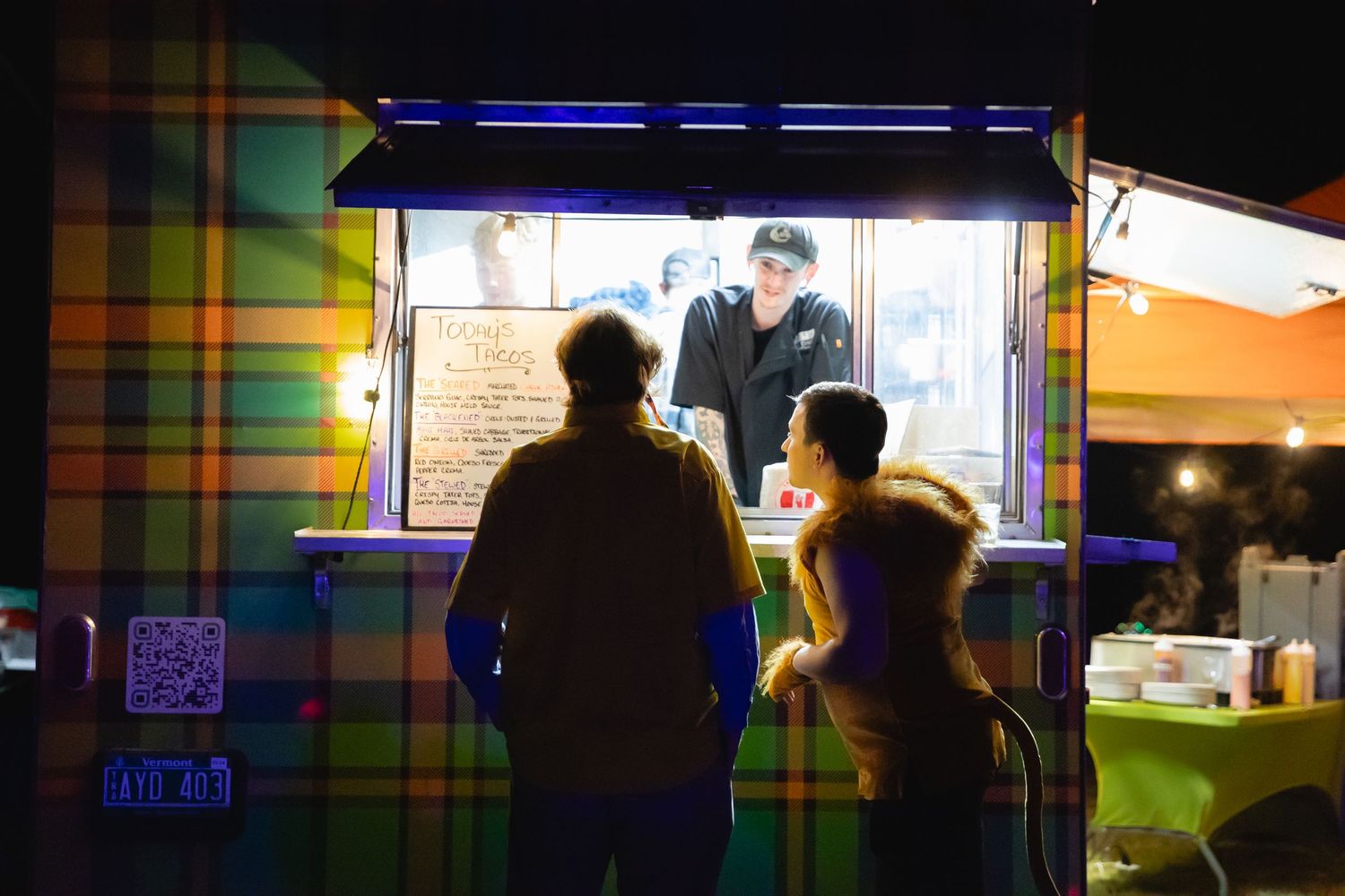 Two customers ordering food from a brightly lit food truck at night.