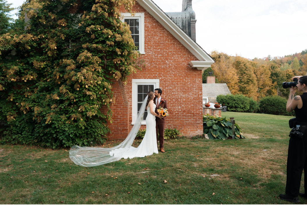 Bride and groom kissing beside brick building with long cathedral veil and autumn foliage.