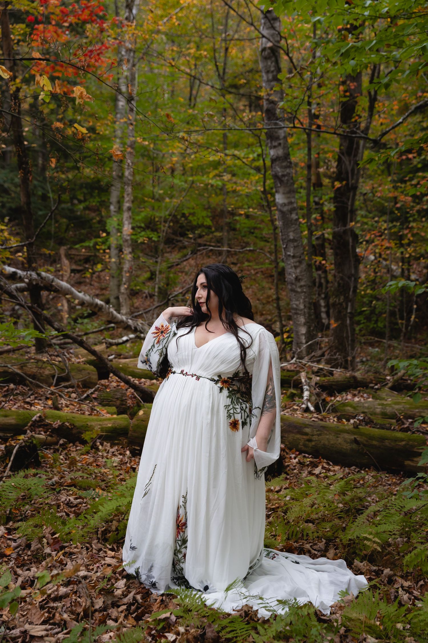 Bride in flowing white dress poses dramatically in autumn forest setting.