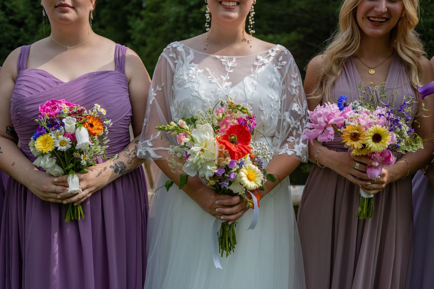 Three bouquets of colorful wildflowers held against purple and white wedding attire.