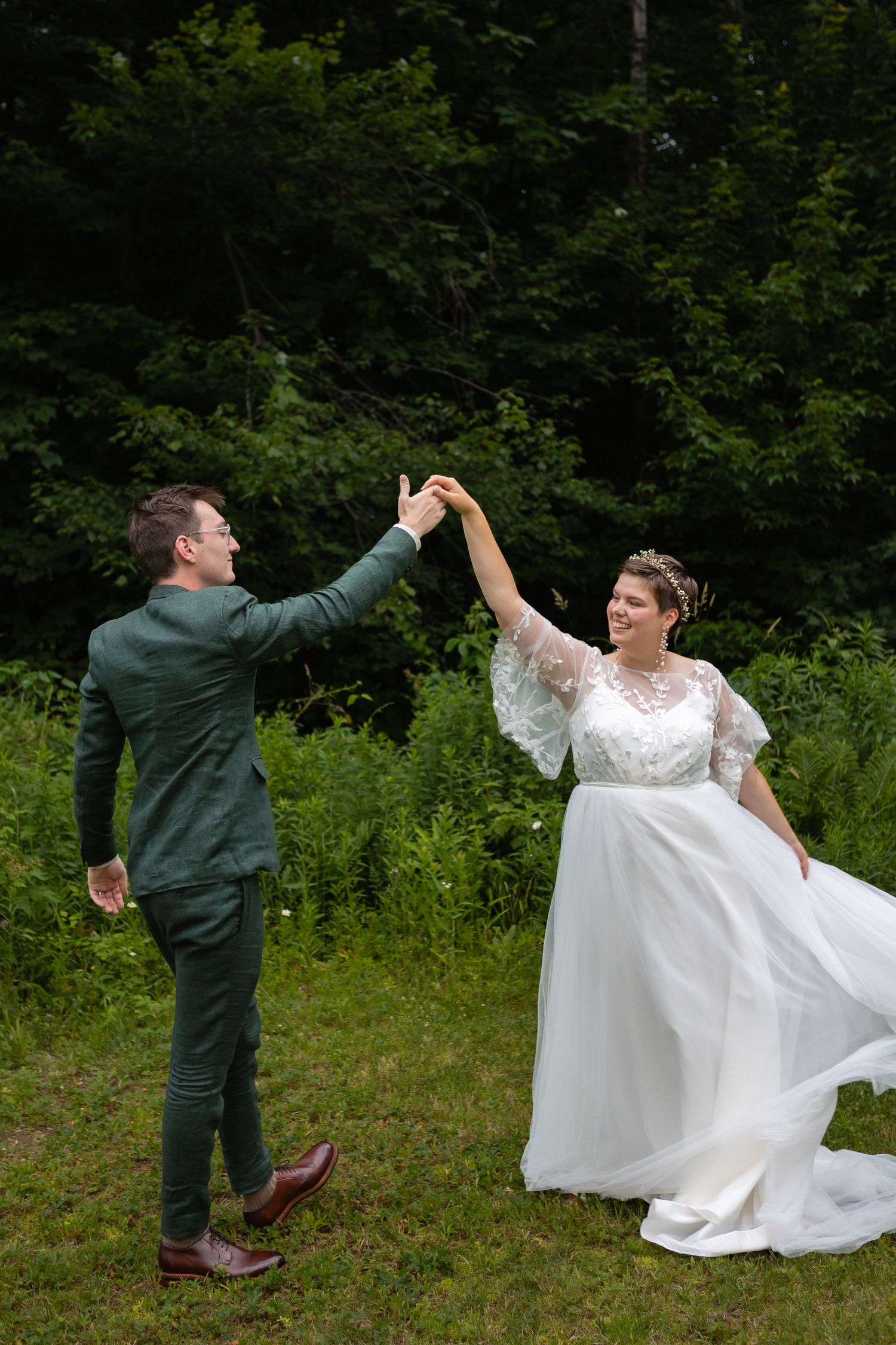 Newlyweds share a joyful dance move on grassy field with forest backdrop.