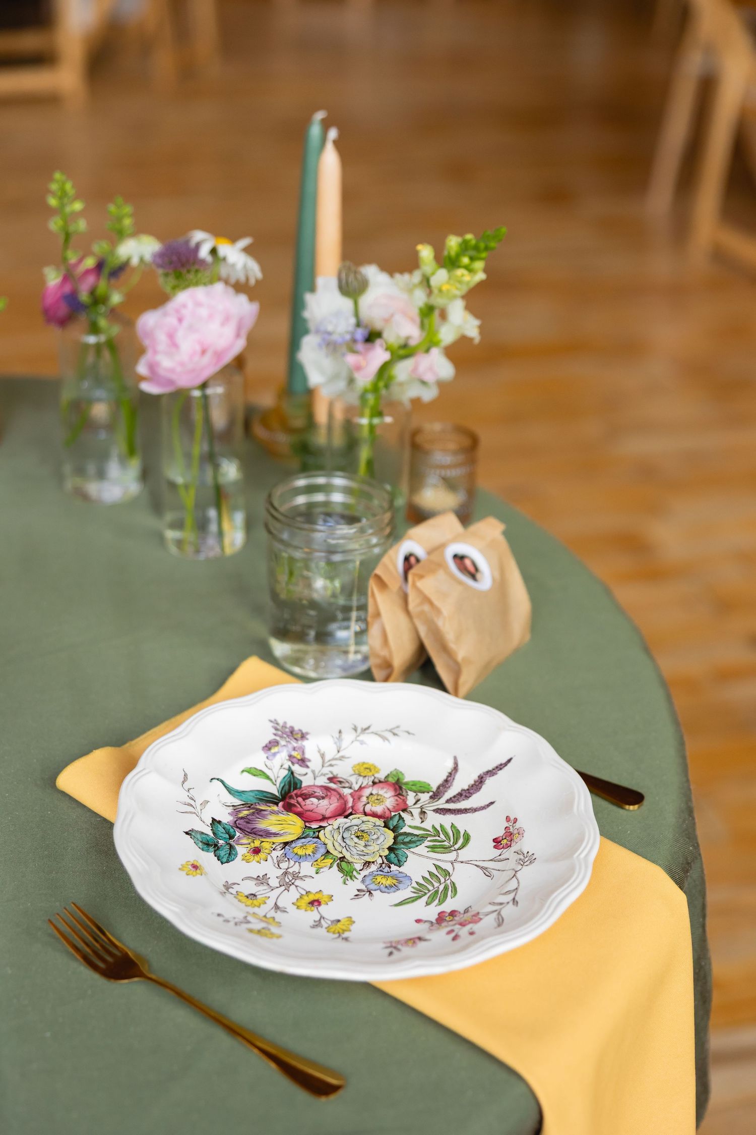 Elegant table setting with floral china plate, yellow napkin, and fresh flowers in glass vases on sage green tablecloth.