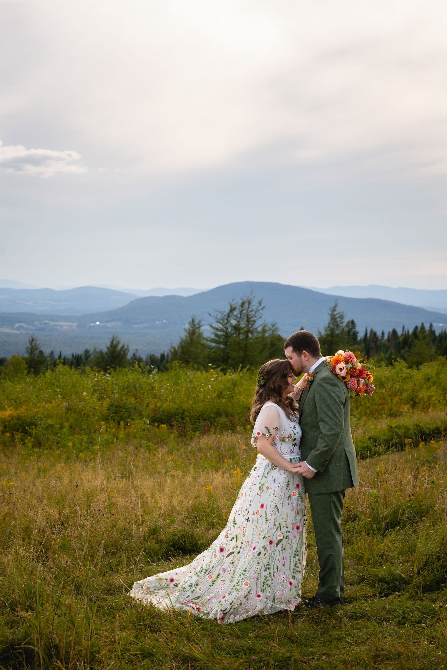 A couple embraces on a hilltop meadow against mountain views, with the bride wearing a floral dress and groom in green suit.