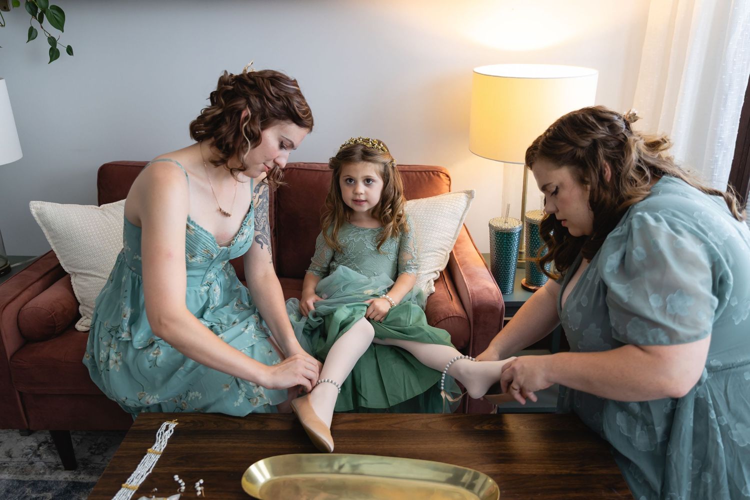 Three people in matching mint green dresses sit on a brown leather couch during a relaxing pedicure session at home.