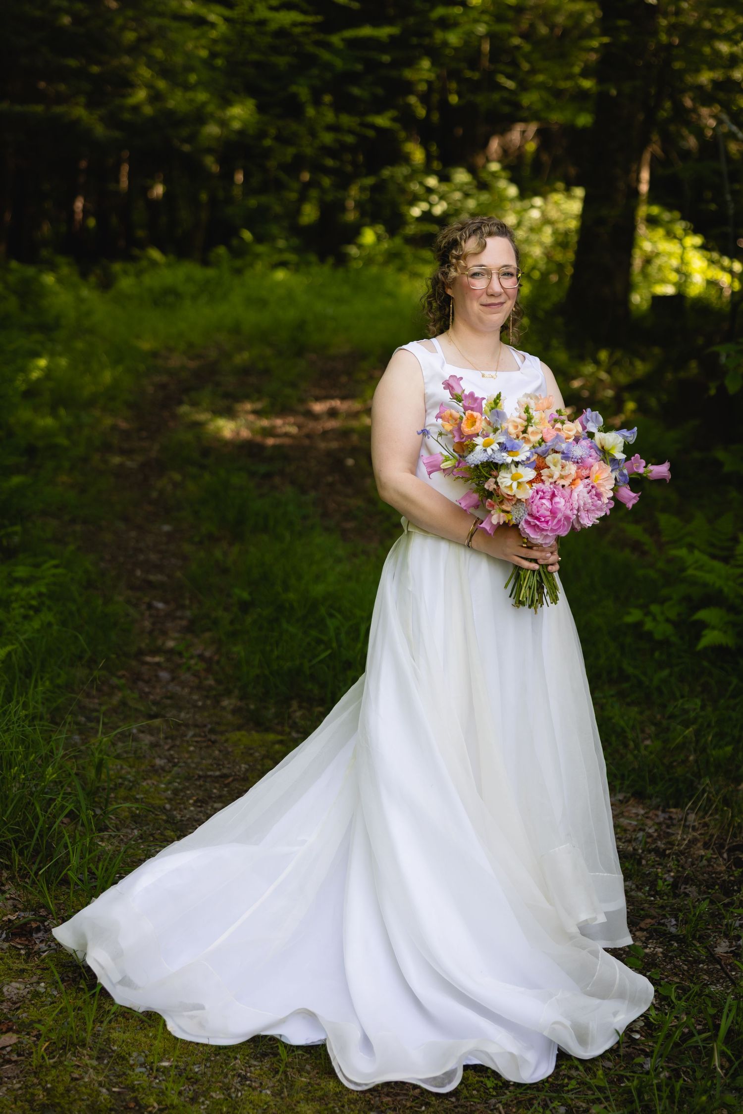 A bride in a flowing white wedding dress holds a vibrant bouquet of pink and purple flowers in a forest setting.