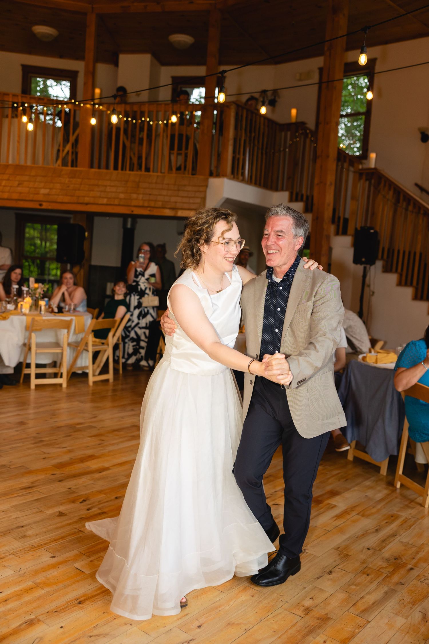 Dancing couple spins on wooden floor at wedding reception with string lights overhead.
