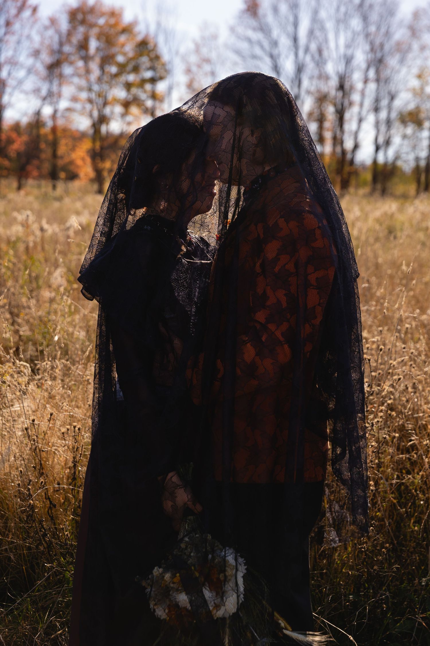 A dark silhouette figure covered in black veil stands in a golden autumn field with fall foliage in the background.
