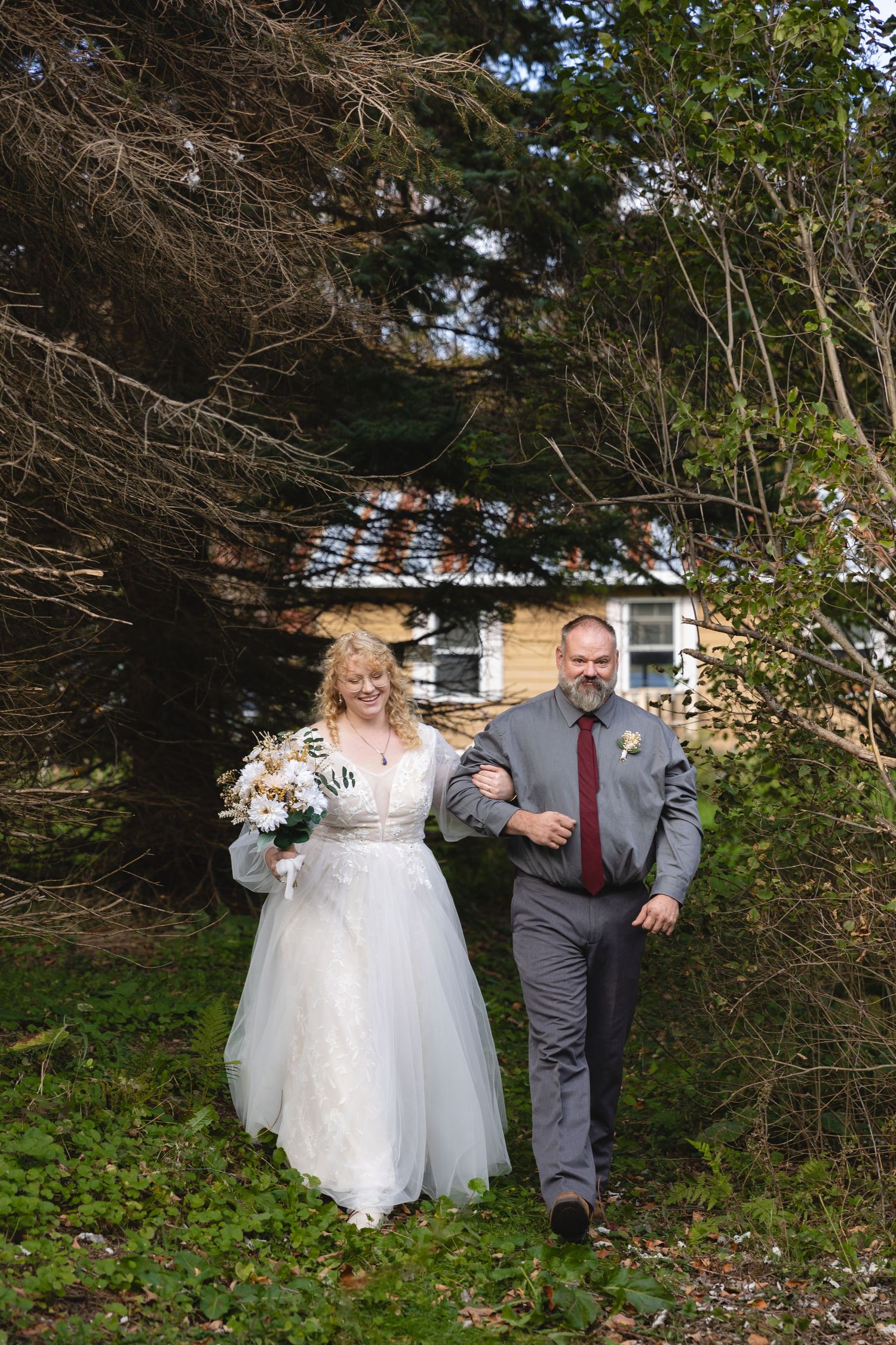 Wedding couple walks down garden path surrounded by trees and greenery.
