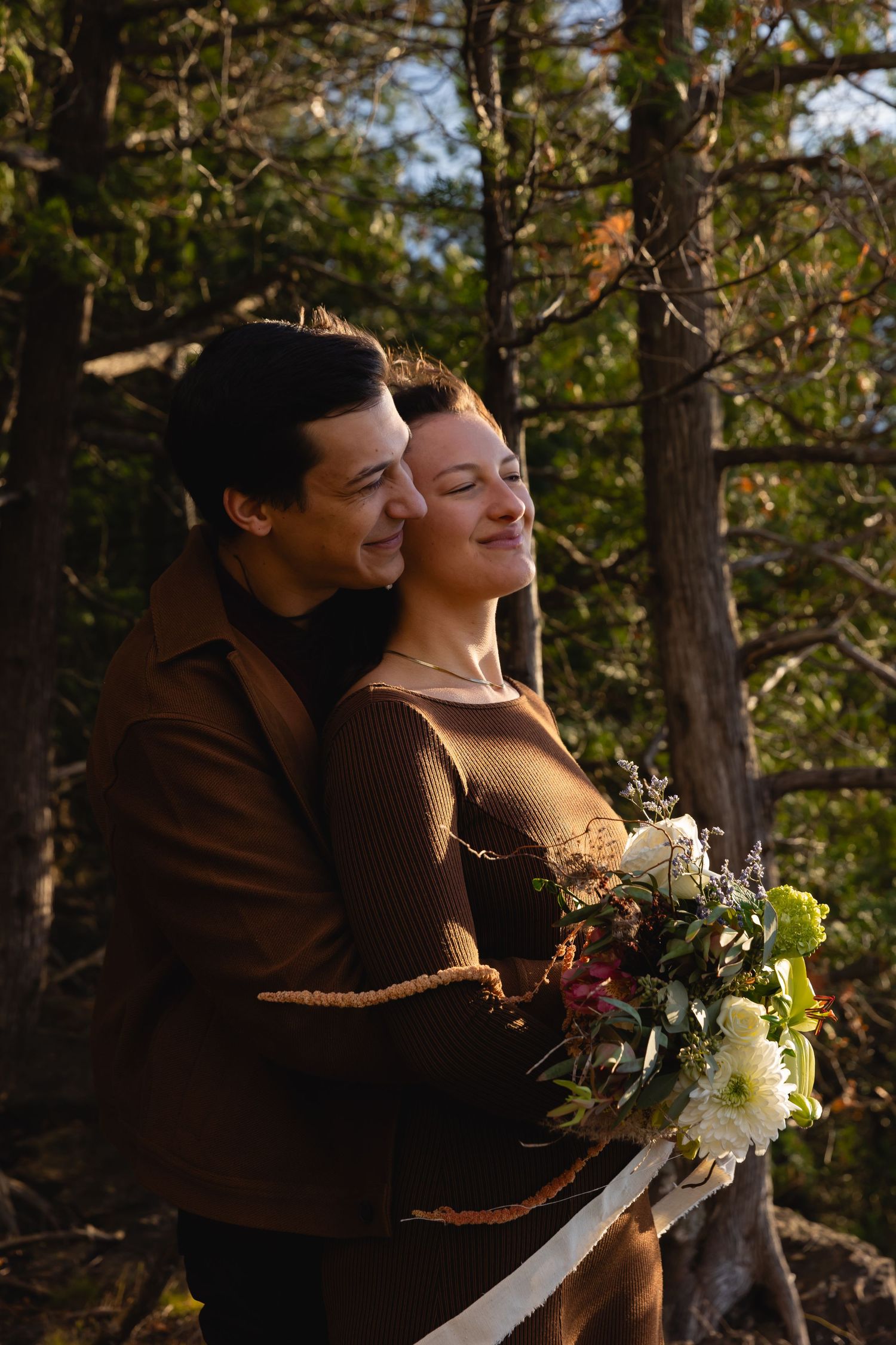 A couple in brown sweaters embrace outdoors in a forest setting with a white floral bouquet.