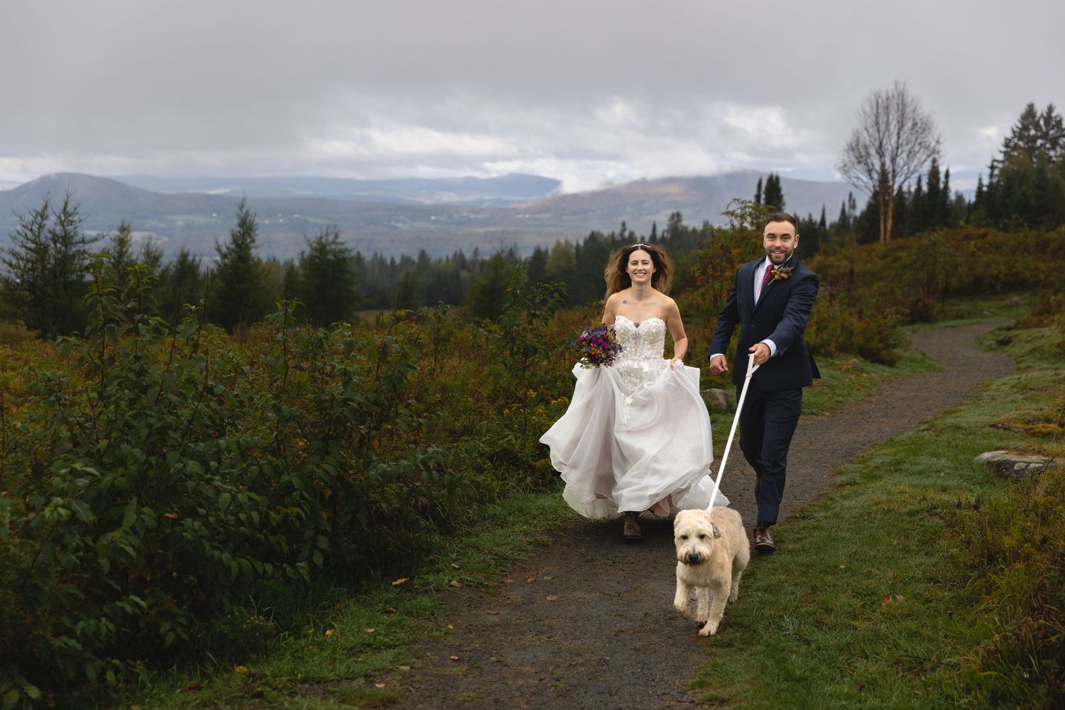 Newlyweds walk their golden dog along a mountain trail surrounded by autumn foliage.