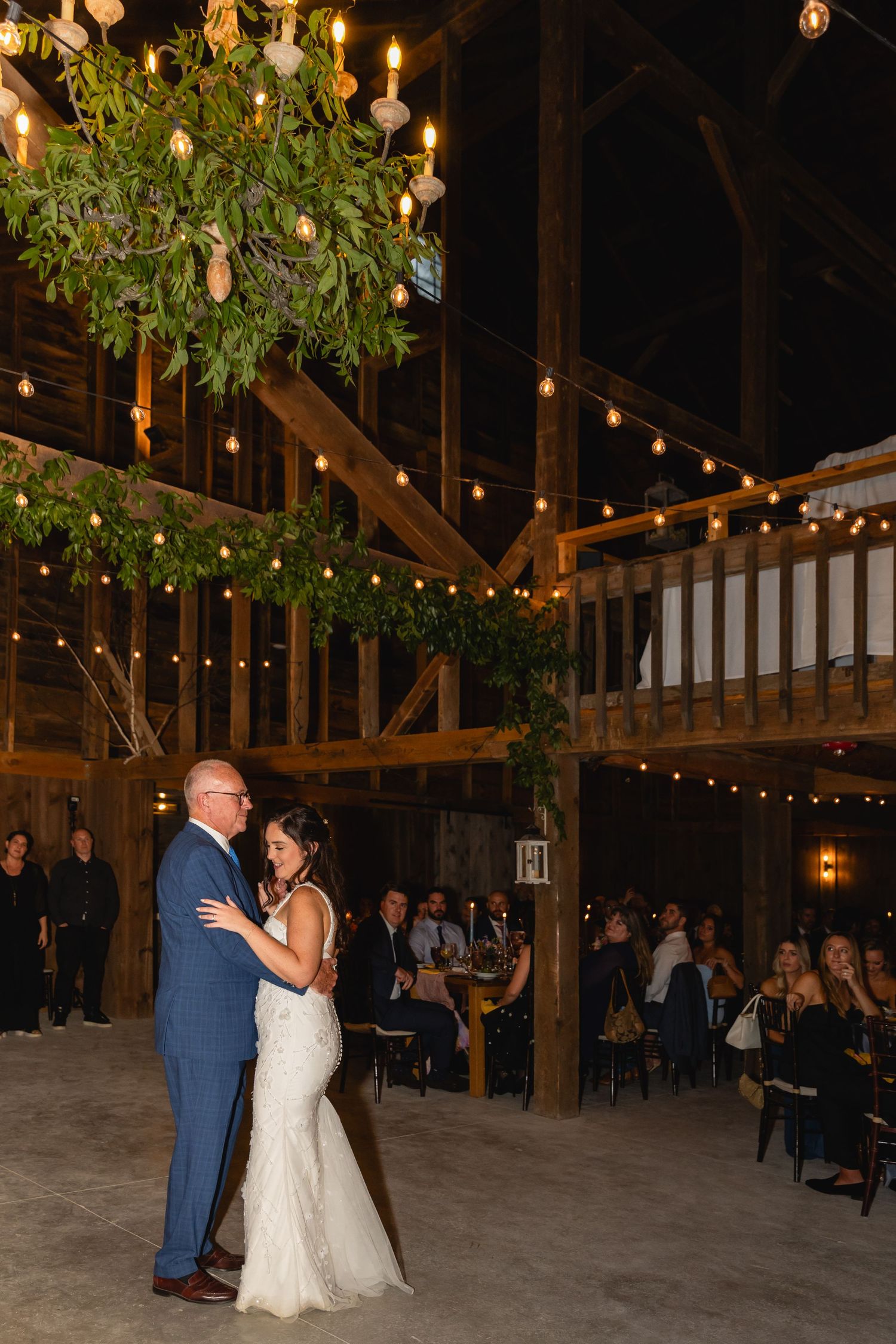 A rustic barn wedding reception with string lights and greenery decorations where two people share a dance on the floor.