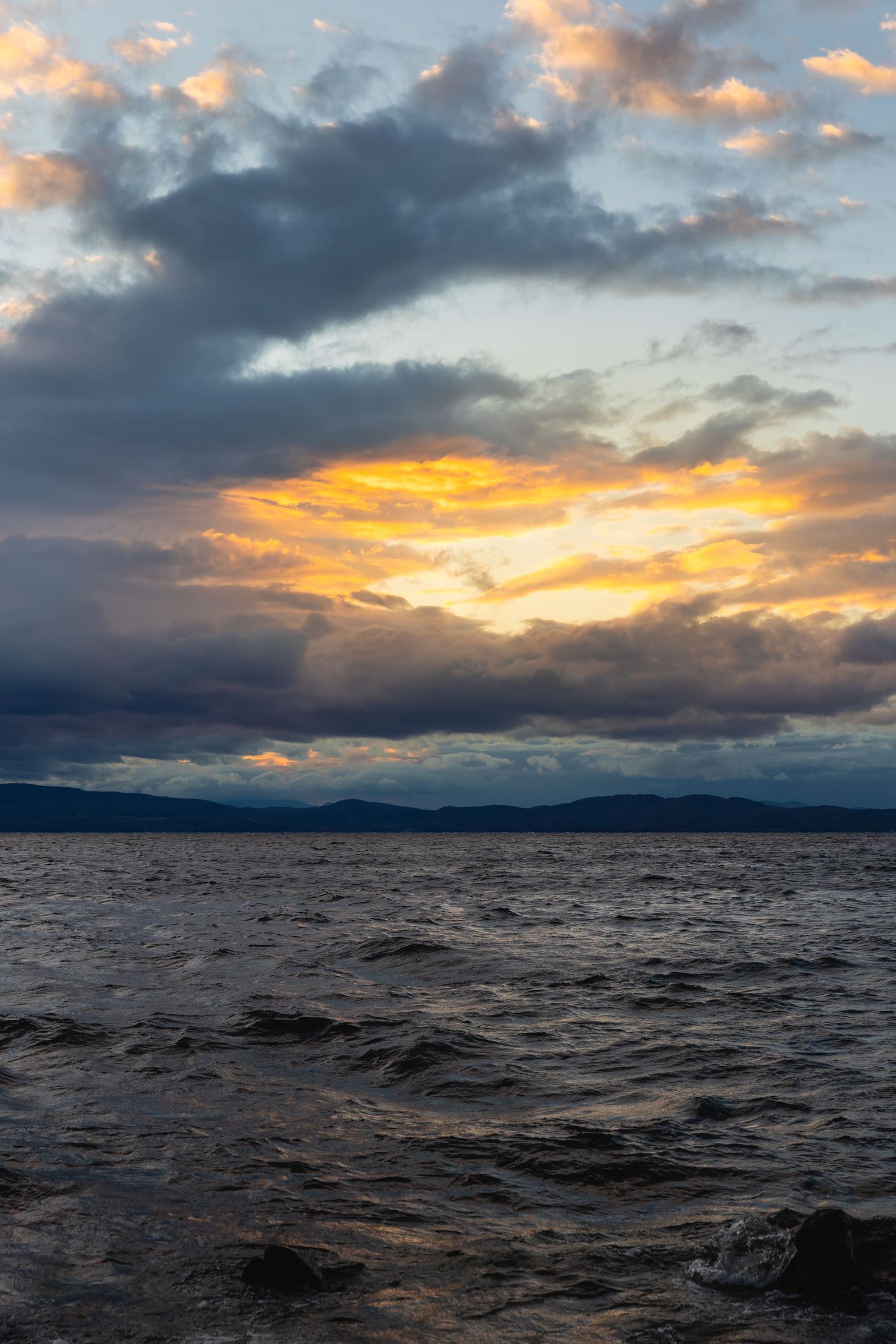 Dark clouds break to reveal vibrant orange sunset over choppy waters of a lake with distant mountains on the horizon.
