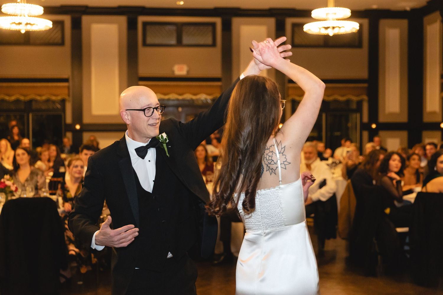 A couple shares a graceful dance moment at an elegant ballroom reception with warm lighting and seated guests watching.