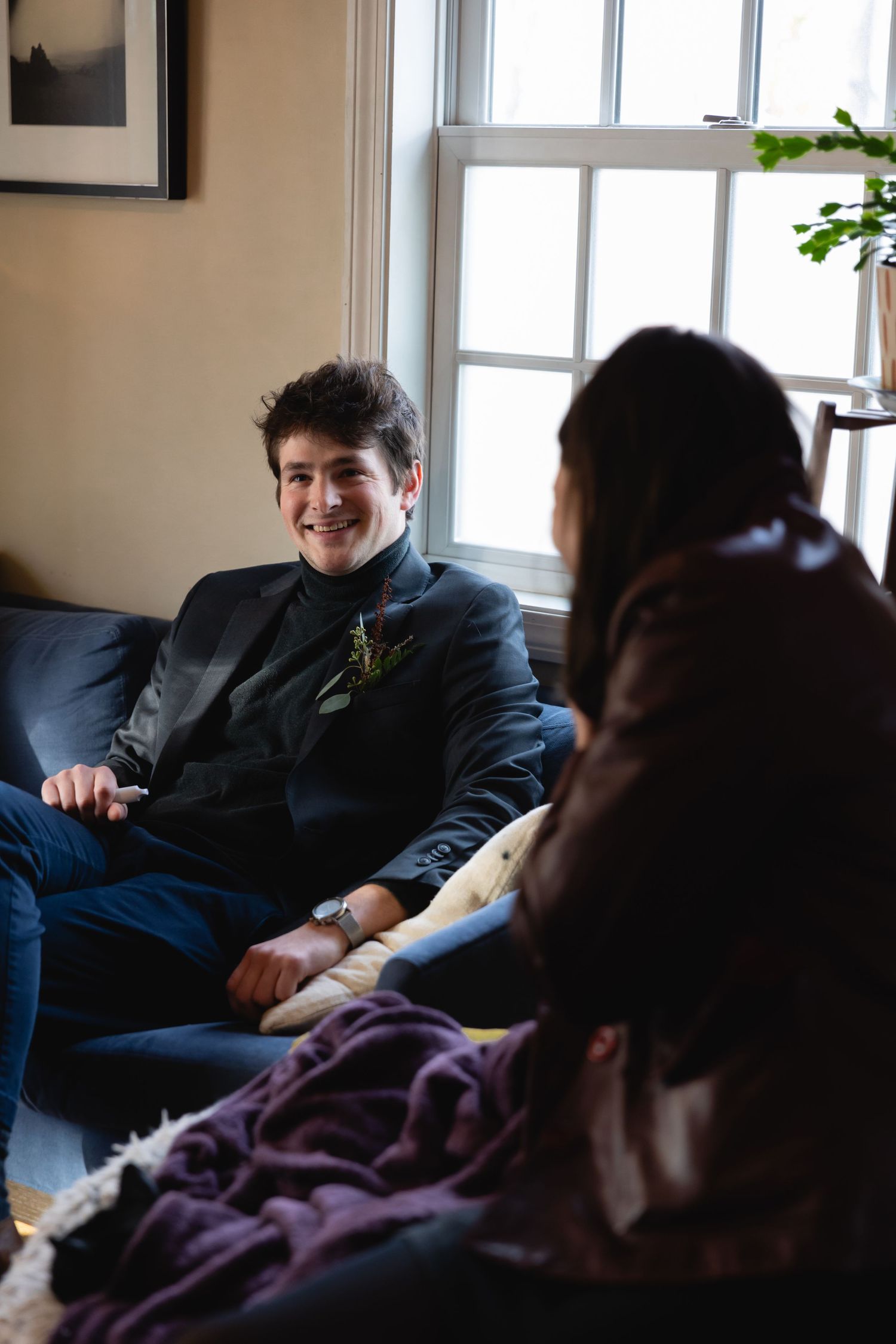 Two people having a relaxed conversation while sitting on a couch by a bright window in a cozy living room.