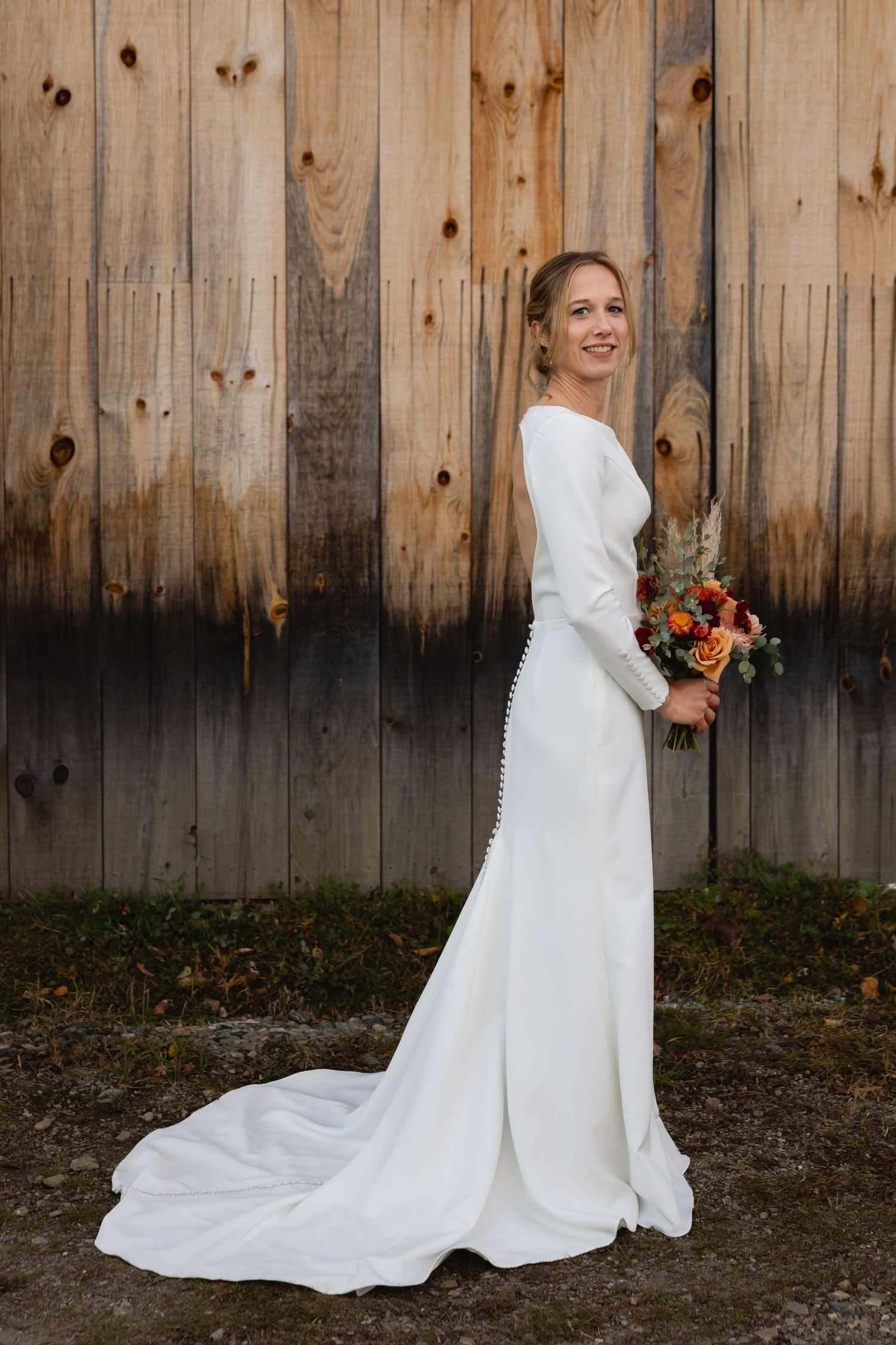 An elegant long-sleeved white wedding dress with a flowing train and fall-colored bouquet against a rustic wooden backdrop.