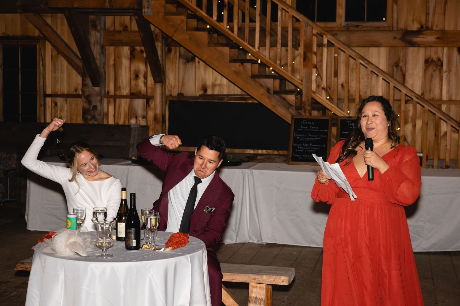 People enjoying a lively moment at a rustic barn reception with someone in red speaking and others seated at a table.