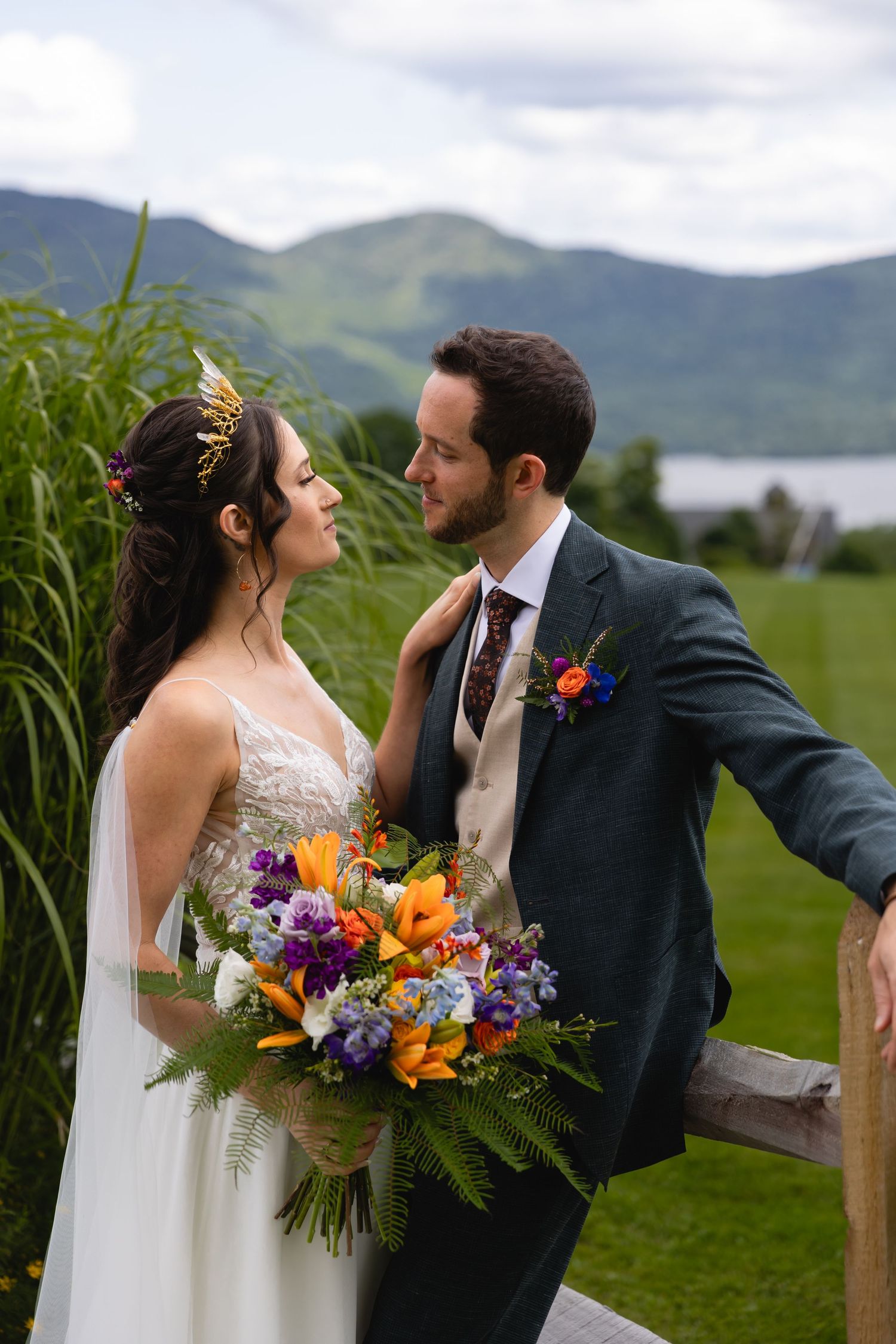 Newlyweds embrace by tall grass with vibrant bouquet against mountain backdrop.