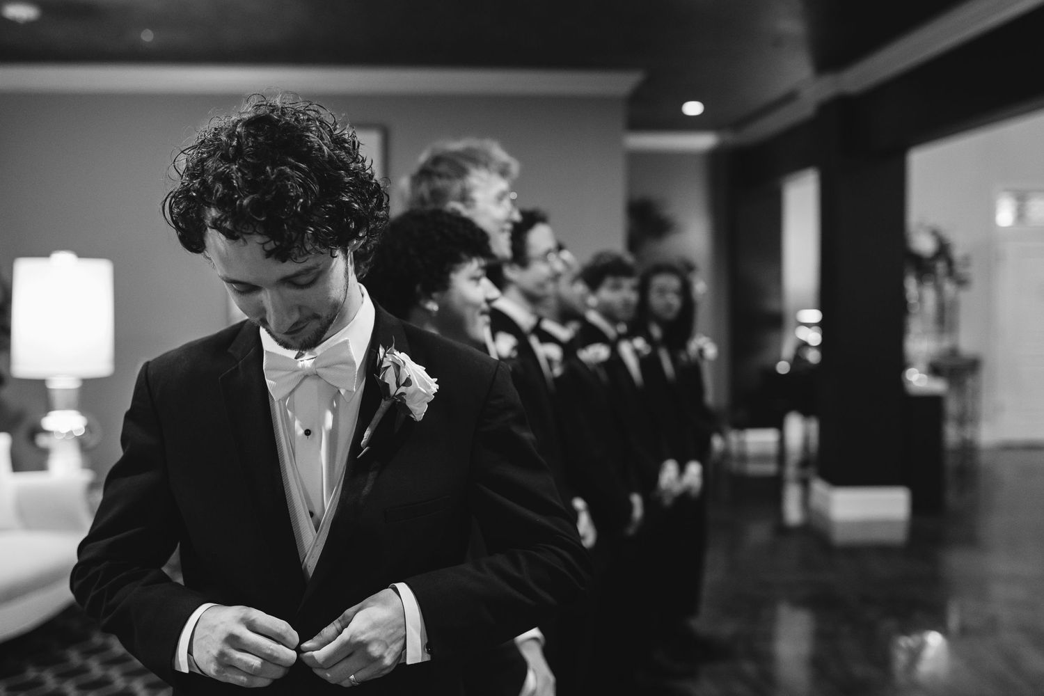A lineup of groomsmen in black and white formal attire with boutonnieres stand in a row at an elegant wedding venue.