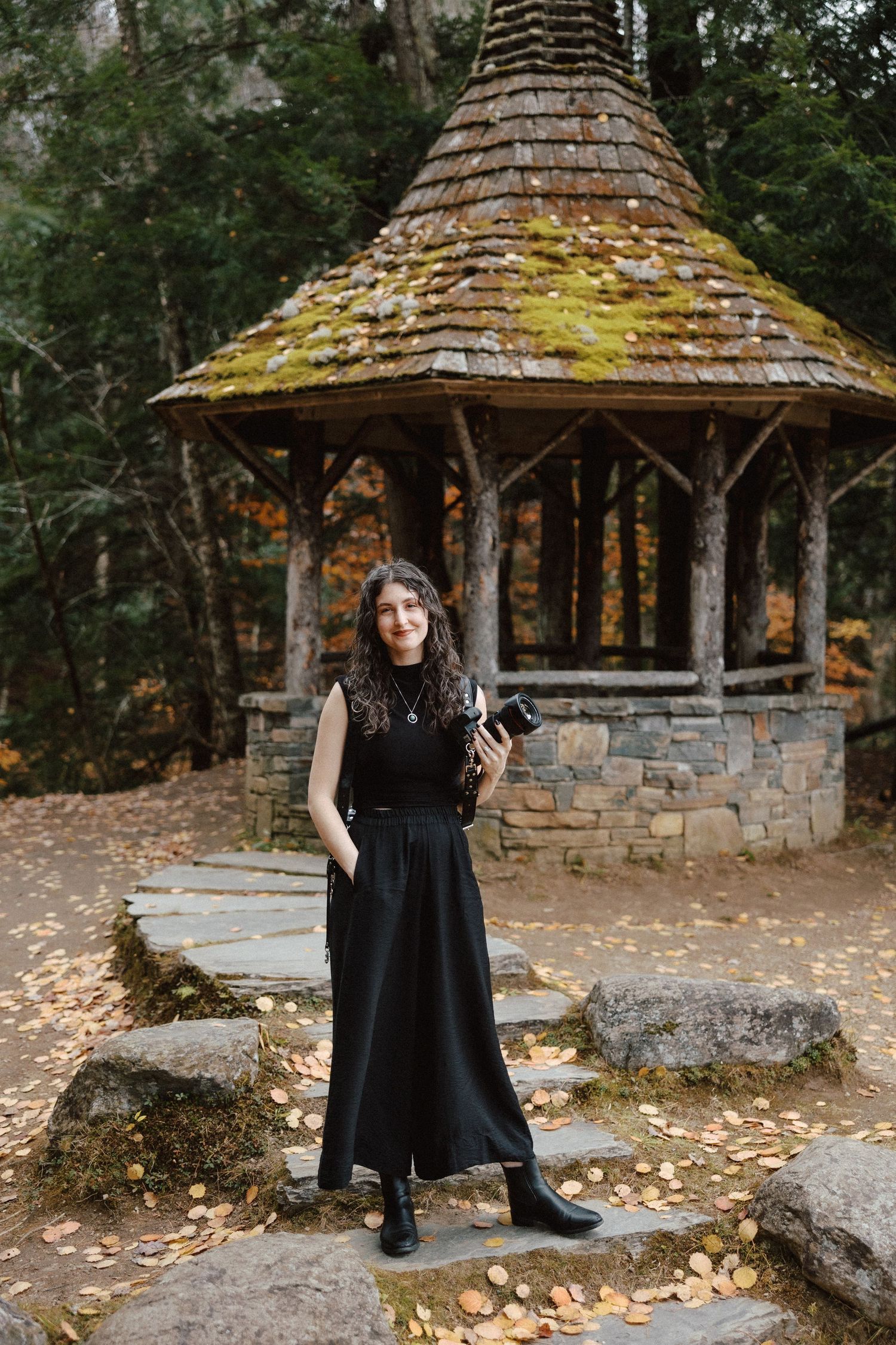 Female photographer in black dress stands before a moss-covered gazebo surrounded by autumn fallen leaves.