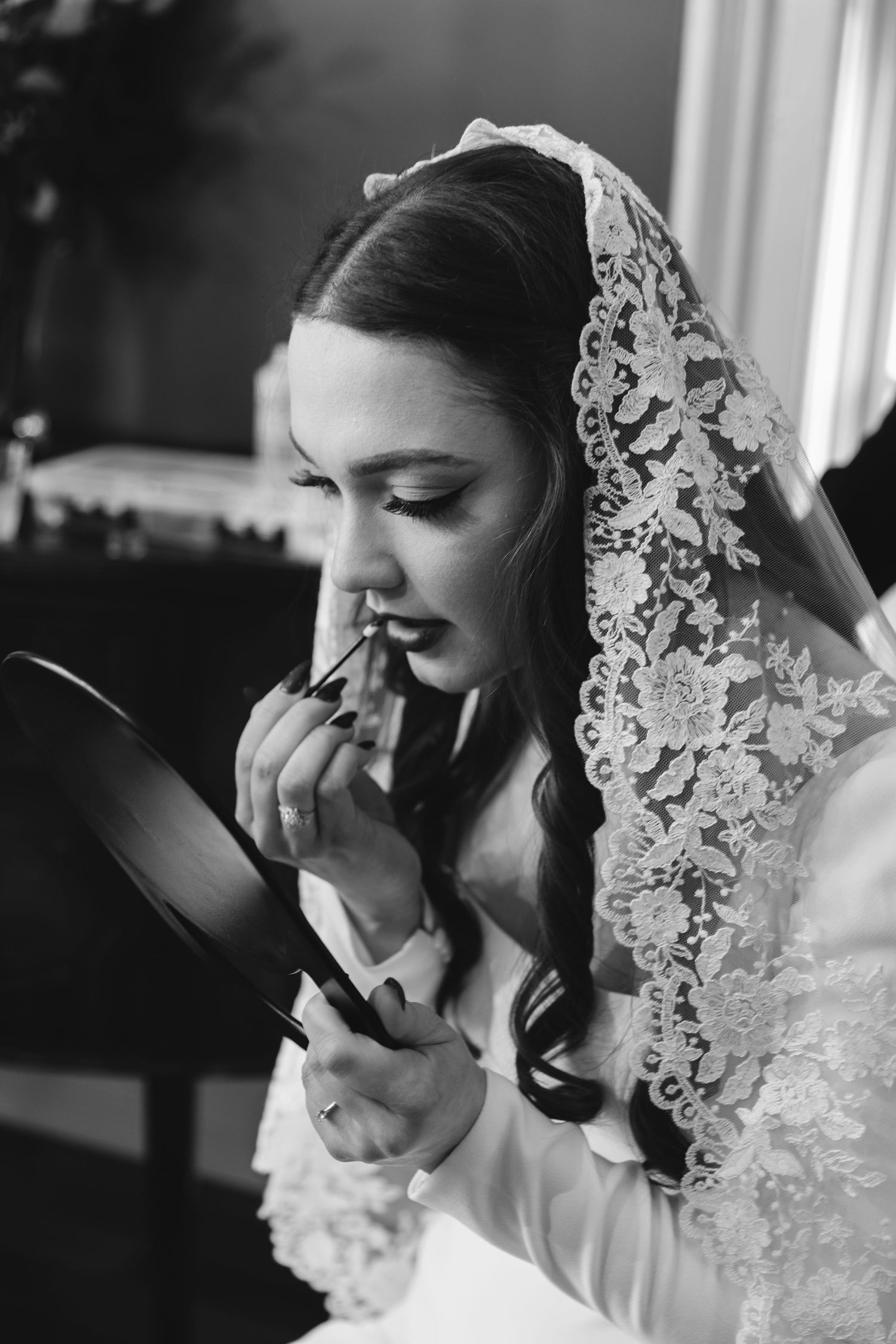 Bride in lace veil applies lipstick using hand mirror in classic black and white bridal preparation photo.