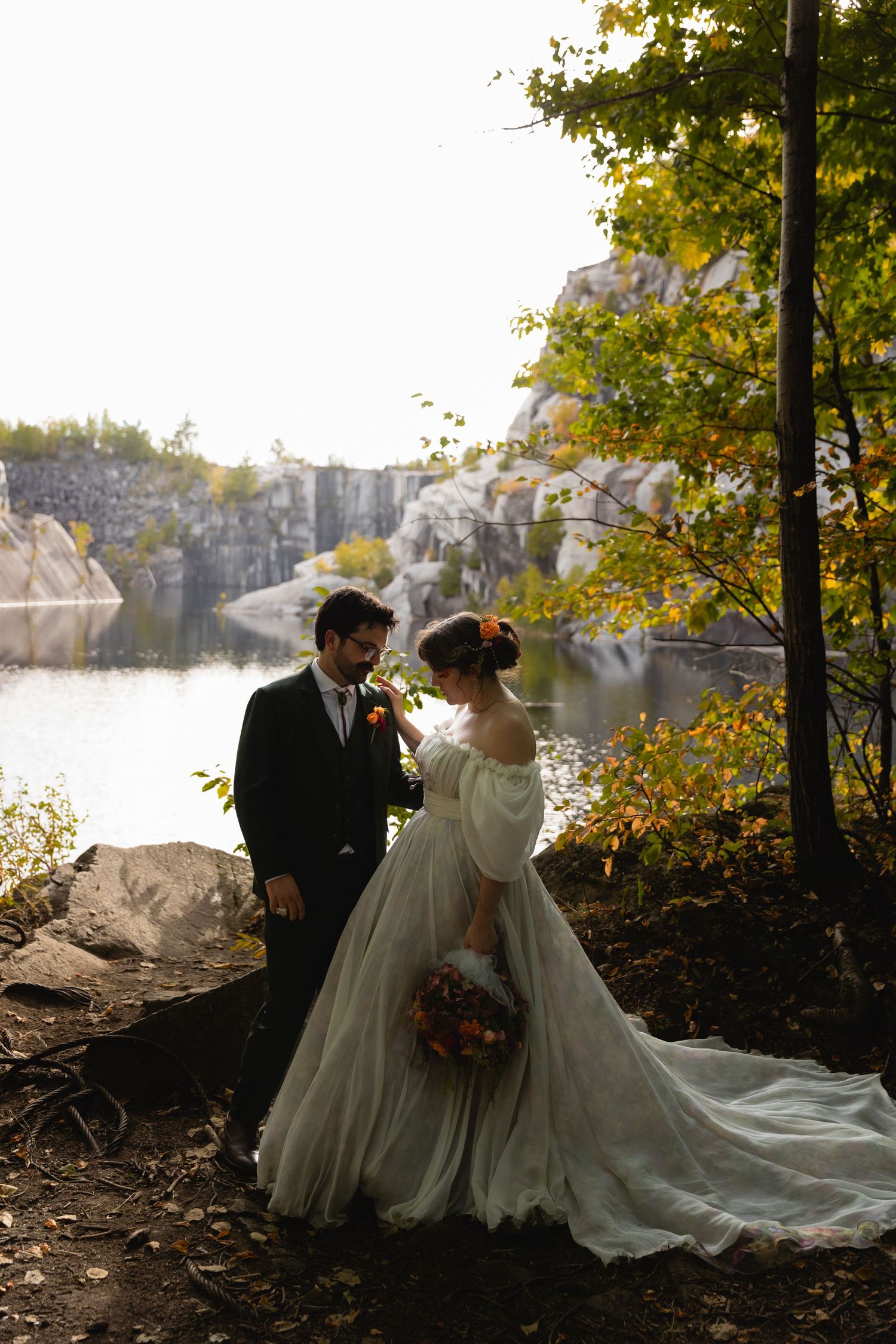 Bride and groom share romantic moment by rocky quarry lake surrounded by fall foliage.