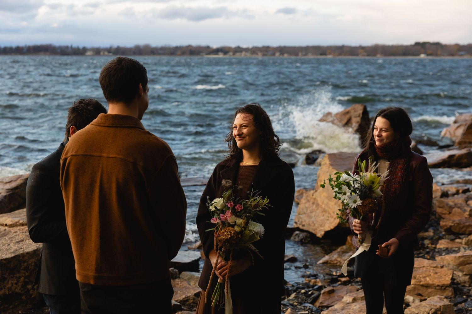 Group of people gathered on rocky shoreline with bouquets, ocean waves crashing behind them.