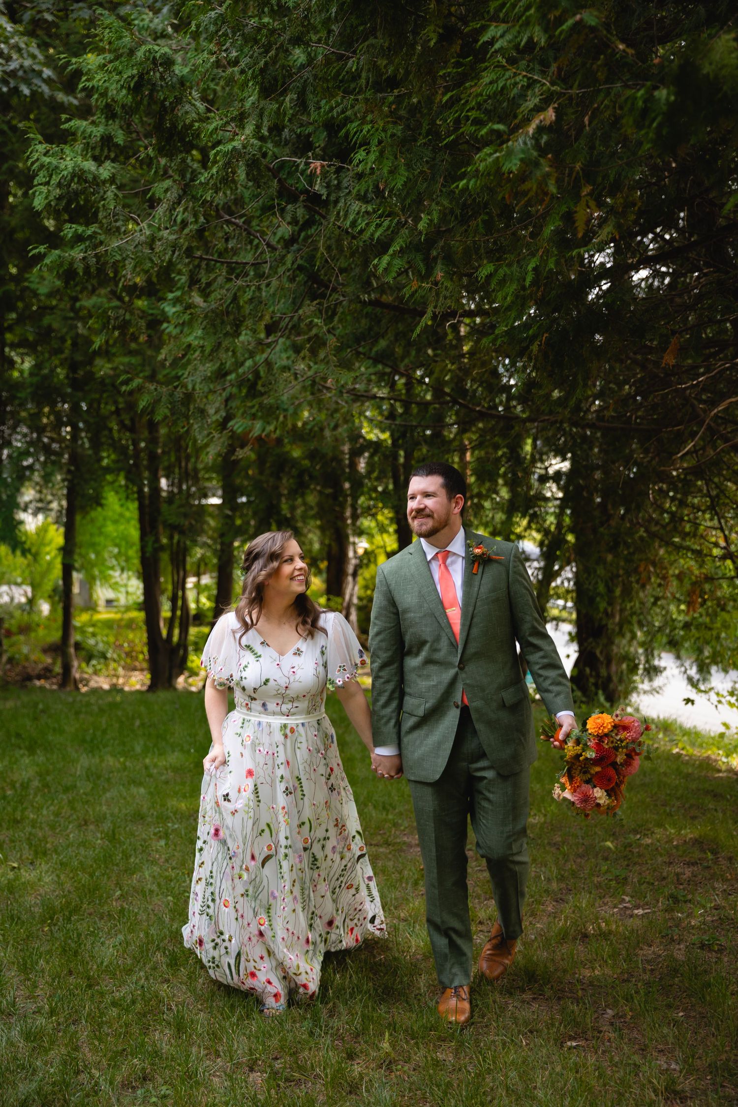 A couple walks through a lush green forest setting wearing a floral dress and green suit for their wedding photos.
