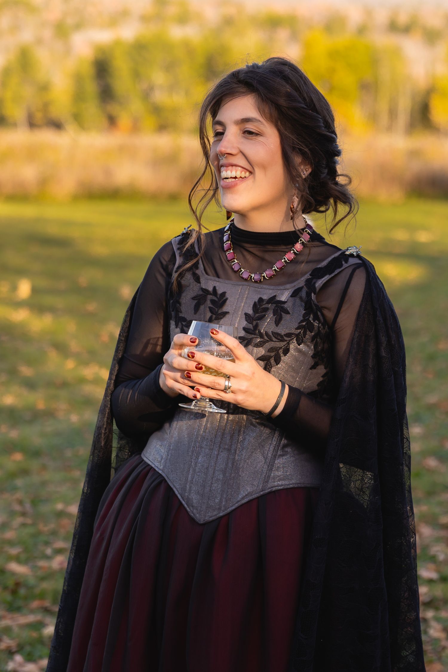 A woman in a dark romantic dress laughs joyfully while holding a cup outdoors during golden hour.