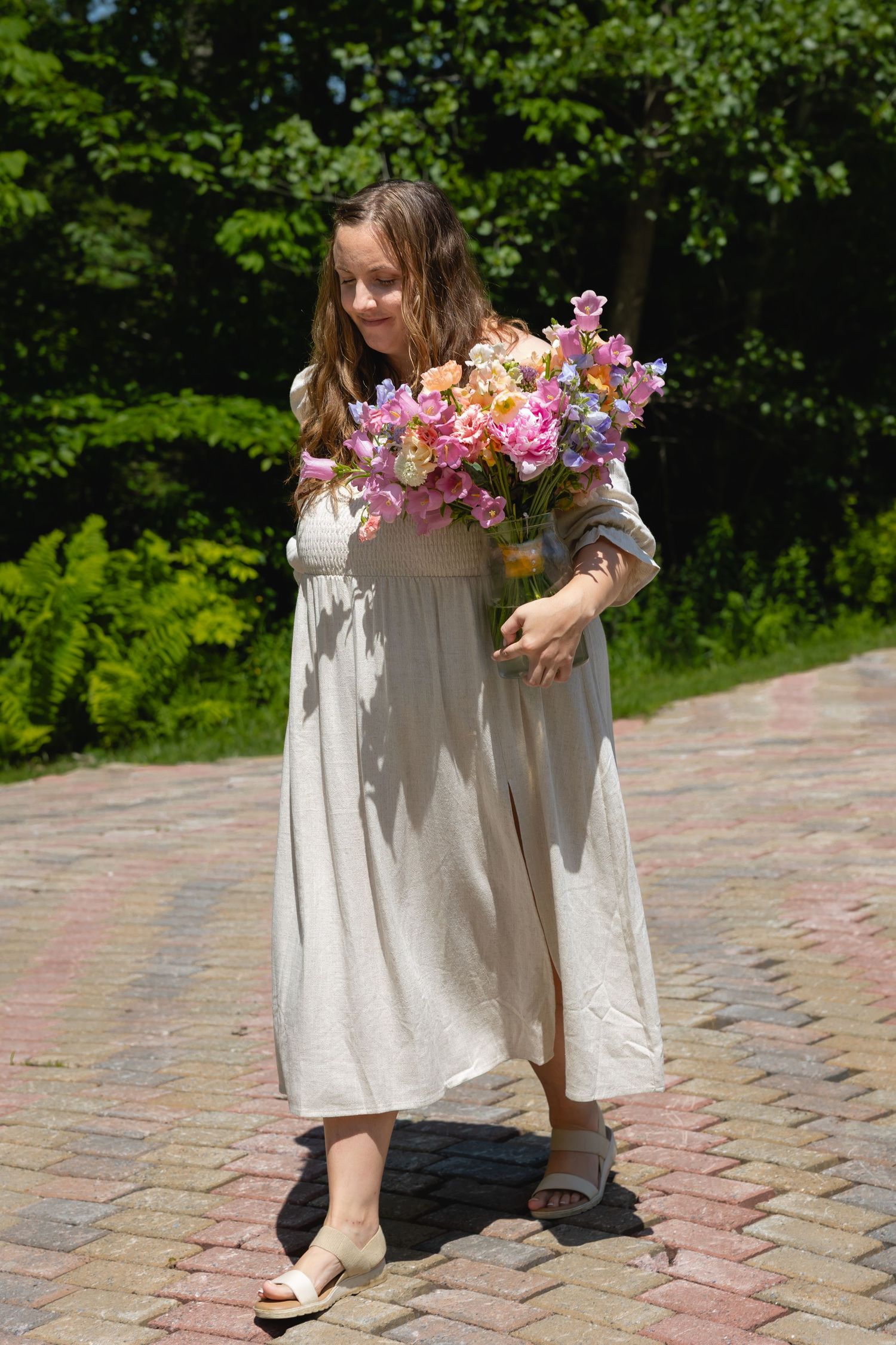 A person in a flowing beige dress stands on a brick path holding a vibrant bouquet of pink and purple flowers.