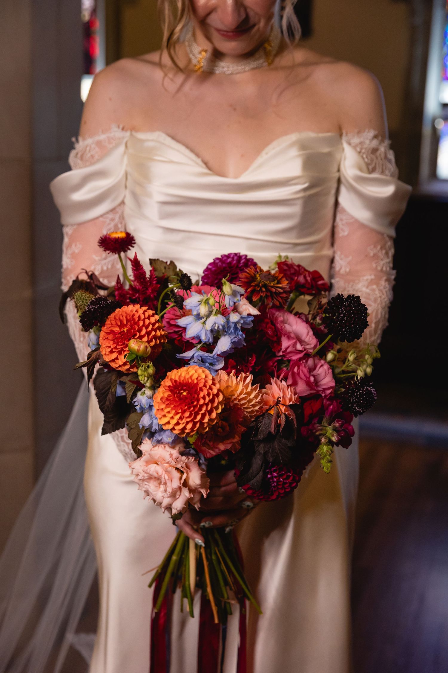 A vibrant autumn wedding bouquet with orange dahlias, burgundy flowers and greenery against a white dress.