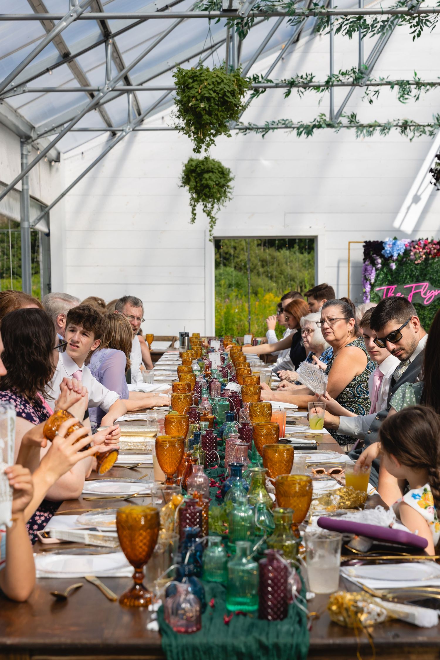 Wedding reception guests seated at a long table in a greenhouse with colorful glass bottles and hanging plants.