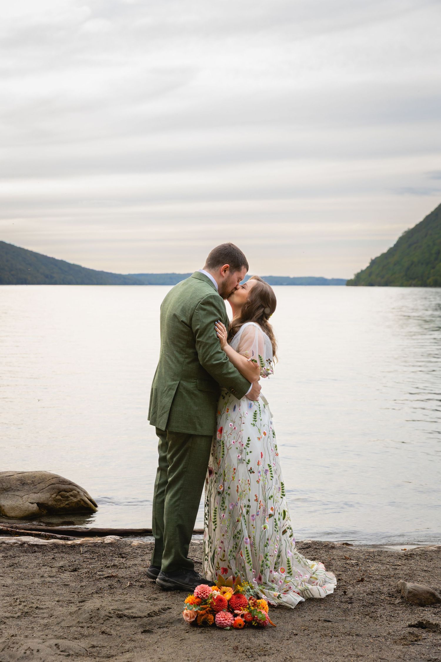 A romantic wedding couple embraces on a rocky beach shore with mountains and water in the background at sunset.