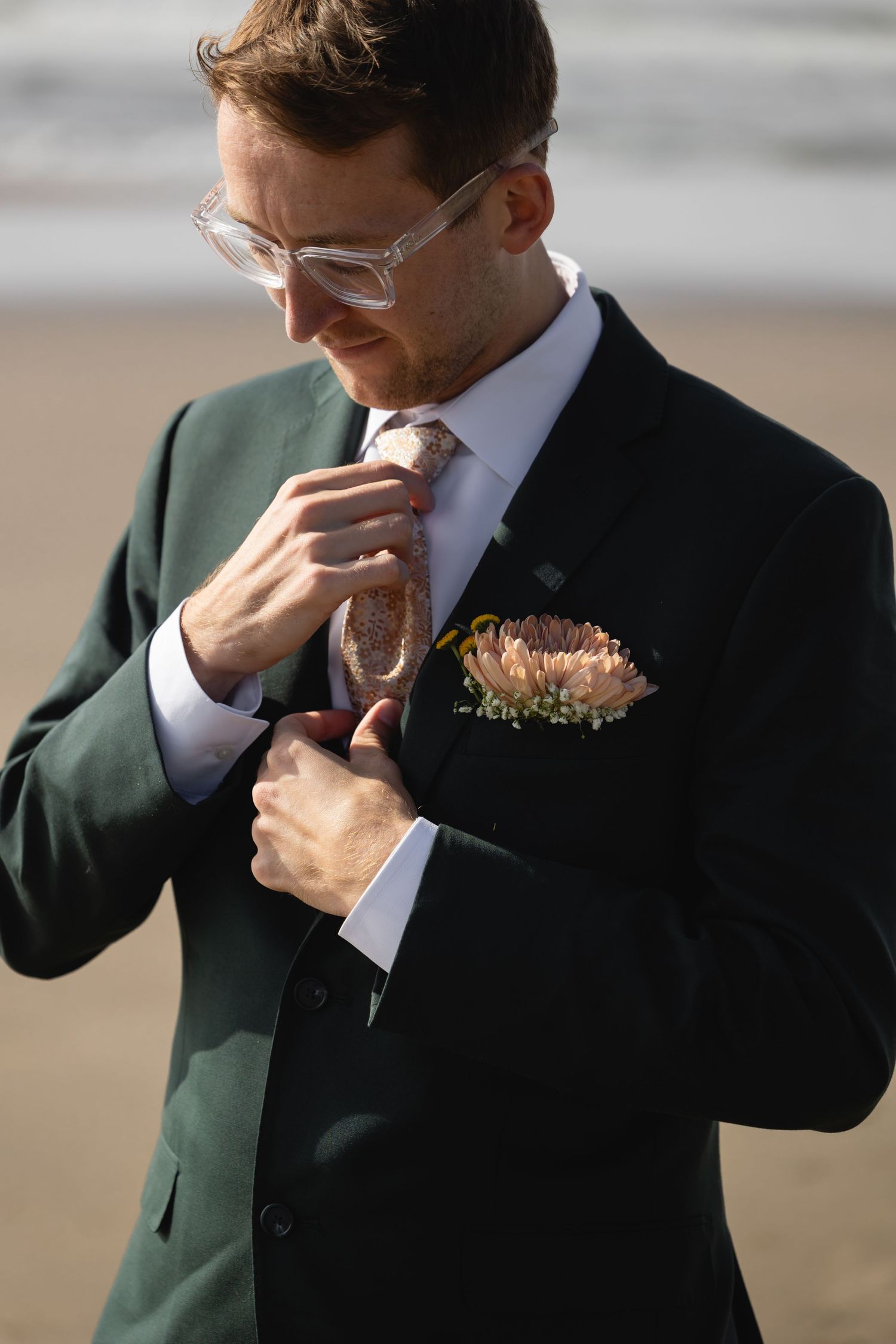 A groom in a dark green suit adjusts his tie while wearing glasses and a floral boutonniere on a beach.