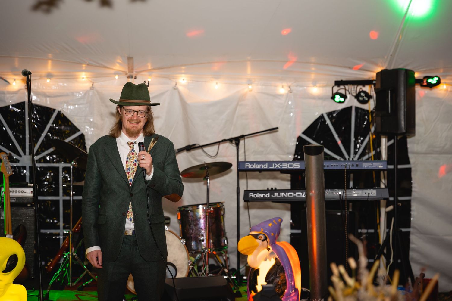 A performer in a green suit and fedora hat stands at a microphone on stage with musical equipment and string lights visible.