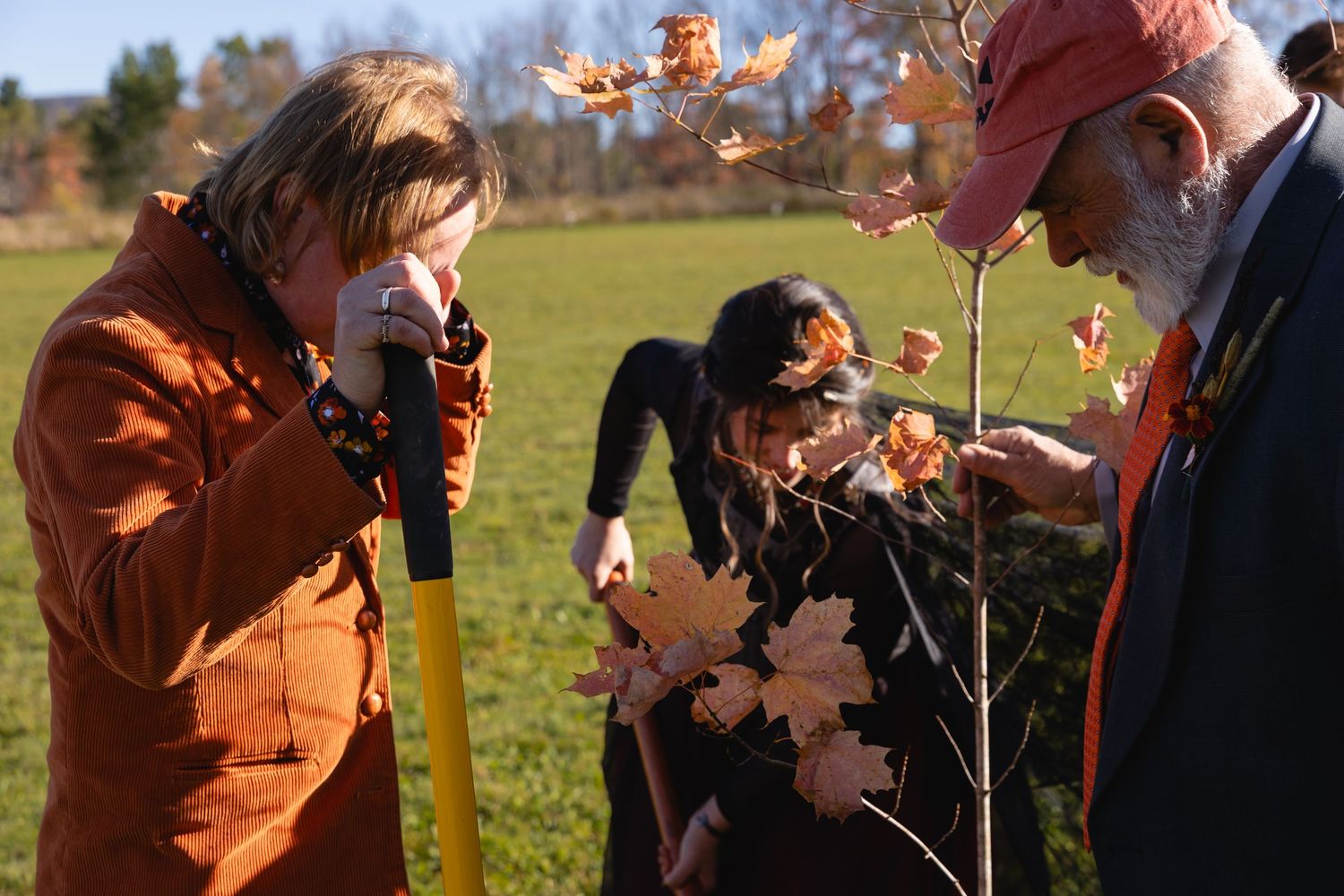 People in orange coats throw autumn leaves in the air while standing in a sunny field on a fall day.